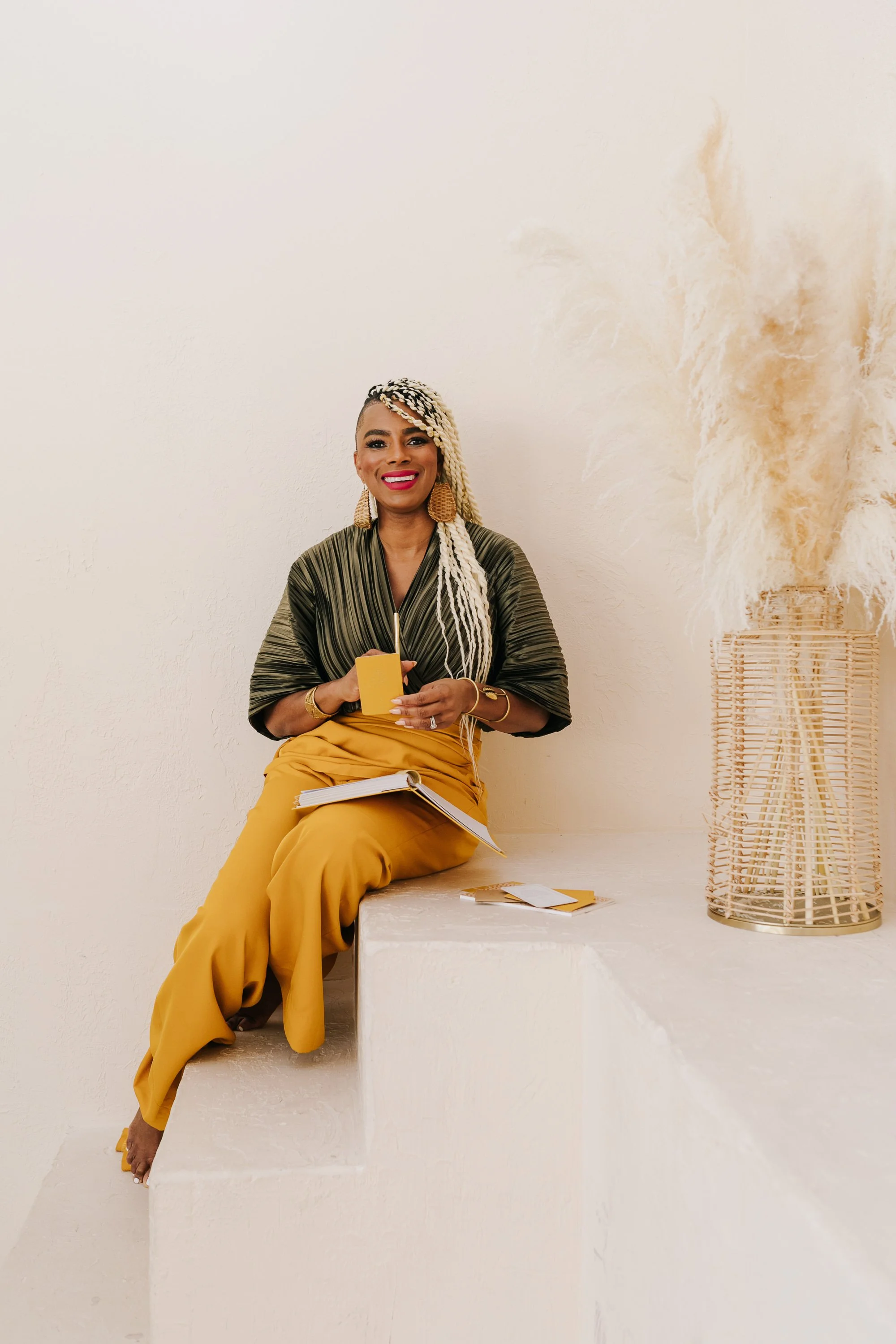 A woman with blonde braids smiling, seated on a white tiered platform, holding a yellow tray, with a notebook on her lap and a yellow paper on the platform, beside a tall decorative vase with pampas grass.