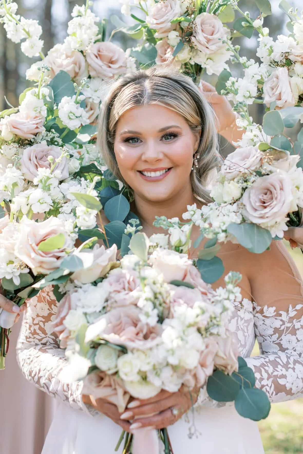 A smiling woman in a wedding dress holding a bouquet of white and blush pink roses and greenery, standing outdoors.