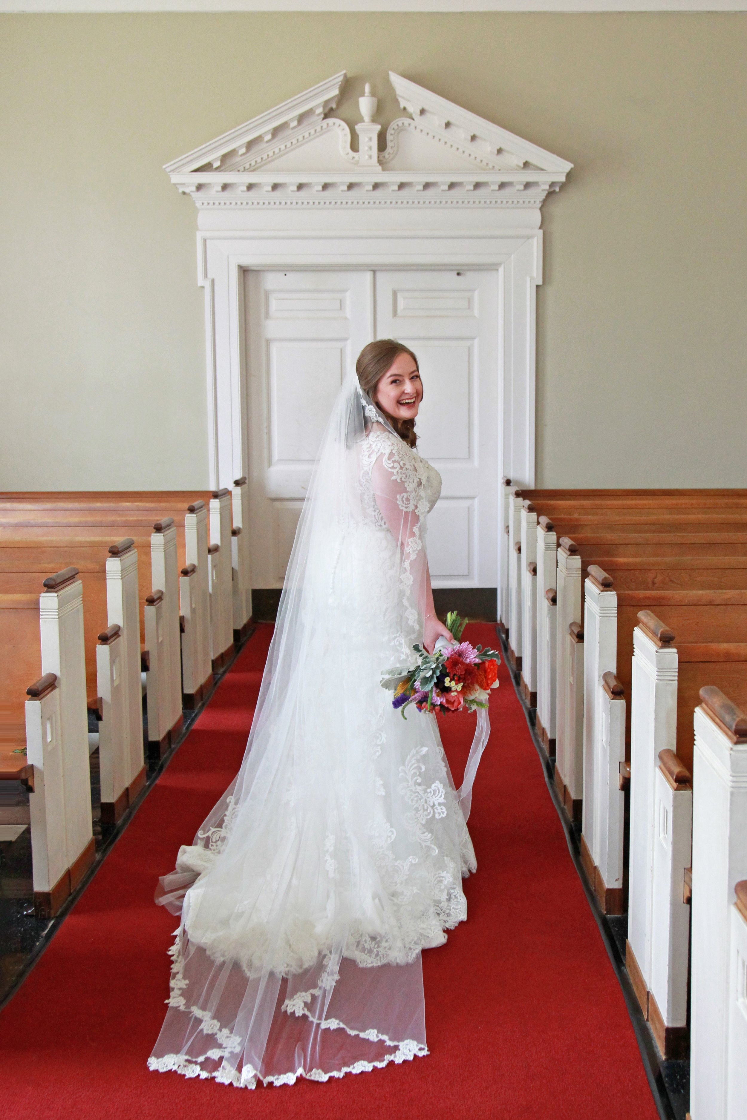 A smiling bride in a white lace wedding dress holding a bouquet of colorful flowers inside a church.