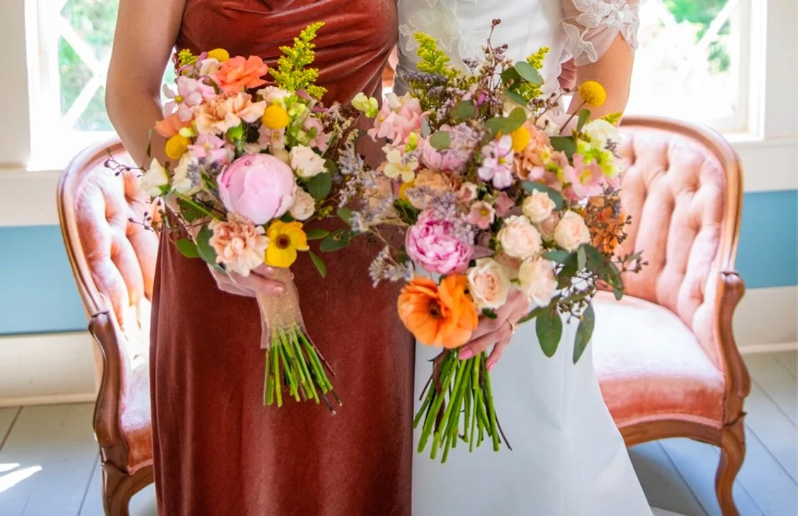 Two women are holding large bouquets of pink, yellow, orange, and purple flowers in a bright room with a pink velvet sofa and windows in the background.