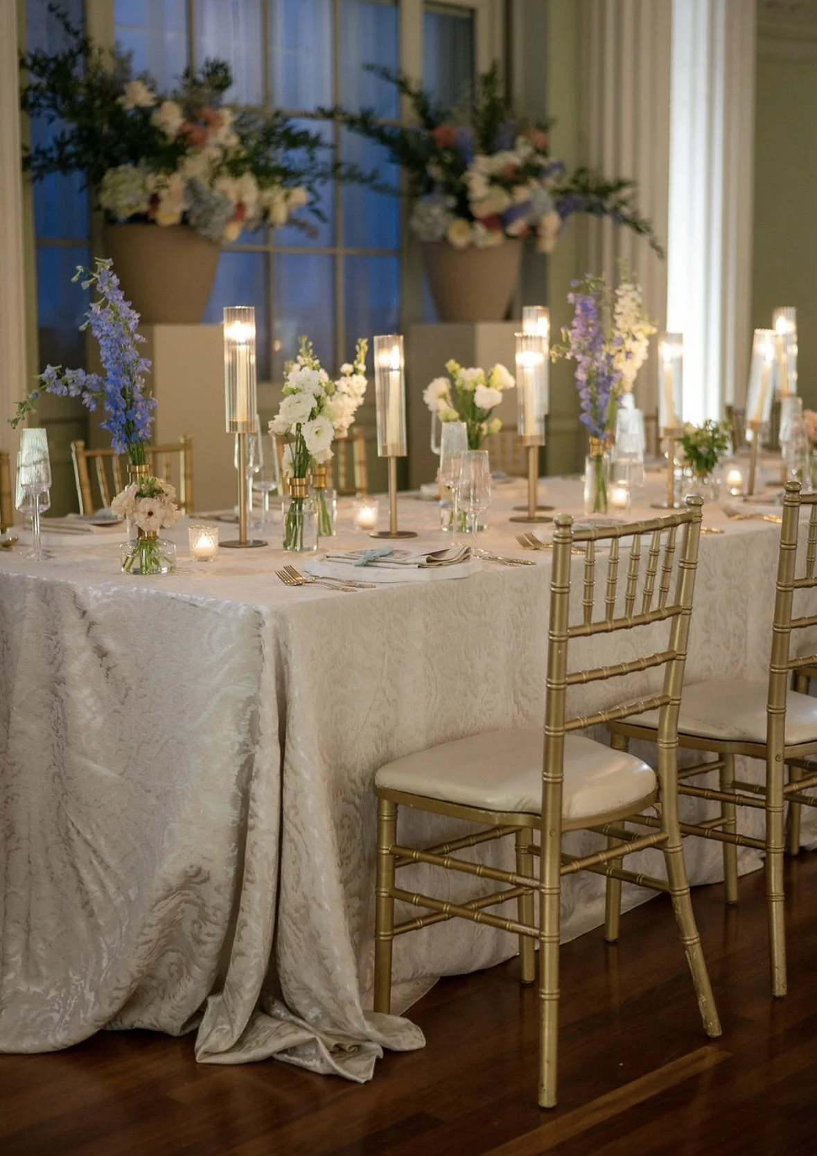 Elegant dining table set with gold chairs, white tablecloth, floral centerpieces, tall candles, and candles in glass holders, in a room with large windows and flower arrangements in the background.