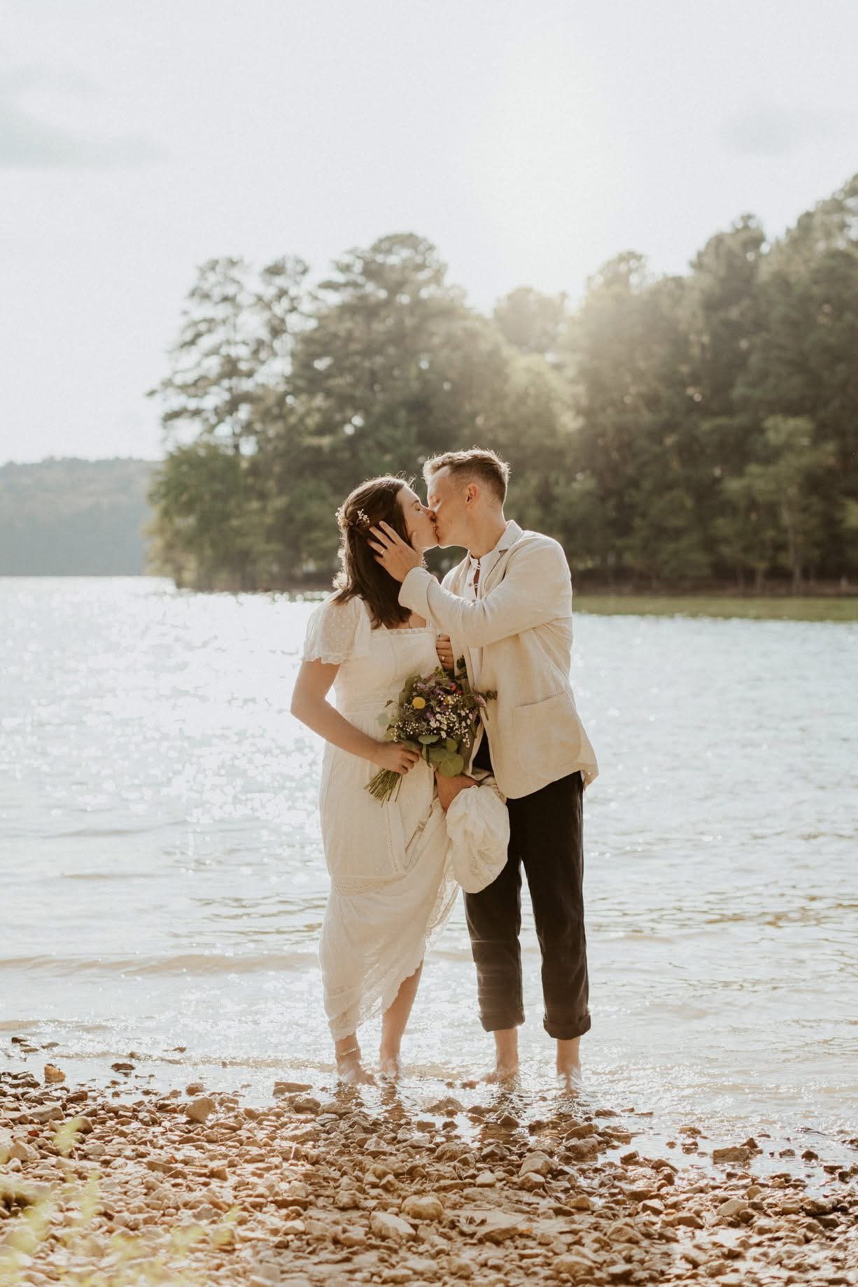 A couple in wedding attire sharing a kiss by a lakeside, holding a bouquet, with trees and sunlight in the background.
