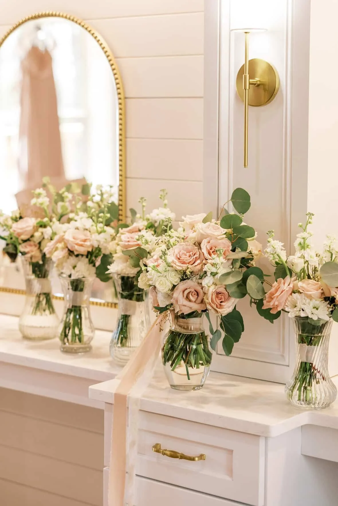Vases with pink and white flowers on a white dresser, reflected in a mirror, with a gold wall sconce and a mirror in the background.