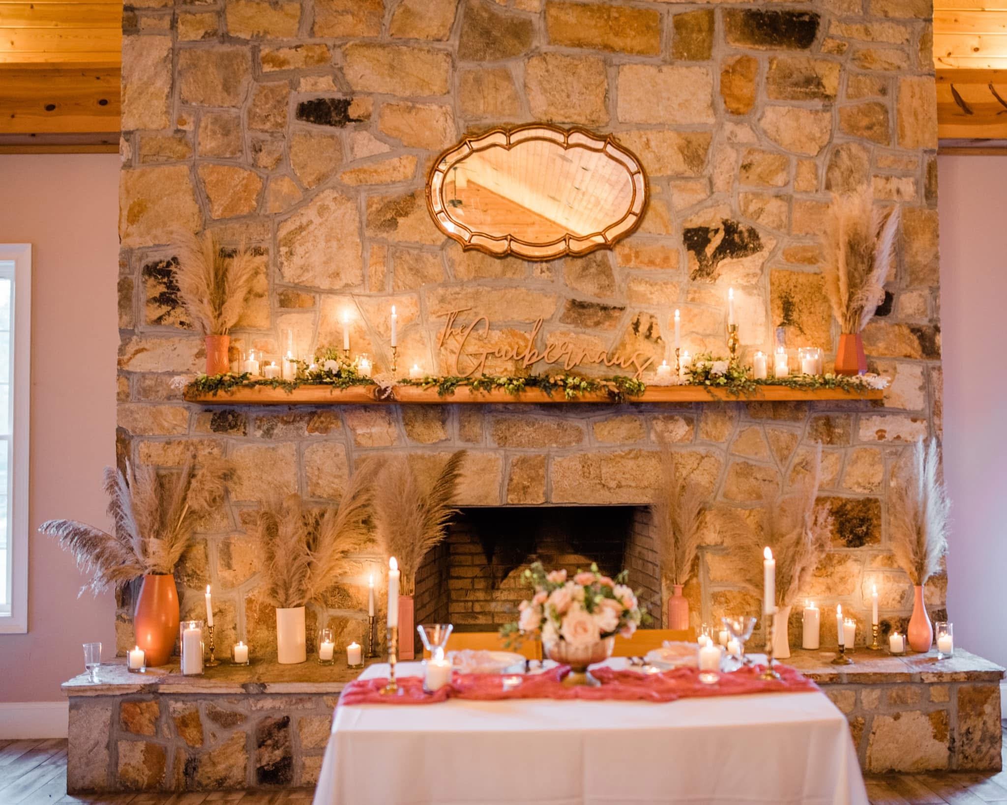A decorated fireplace mantel with candles, pampas grass in vases, and a 'The Gathering' sign, with a table in front set with flowers, candles, and glassware.