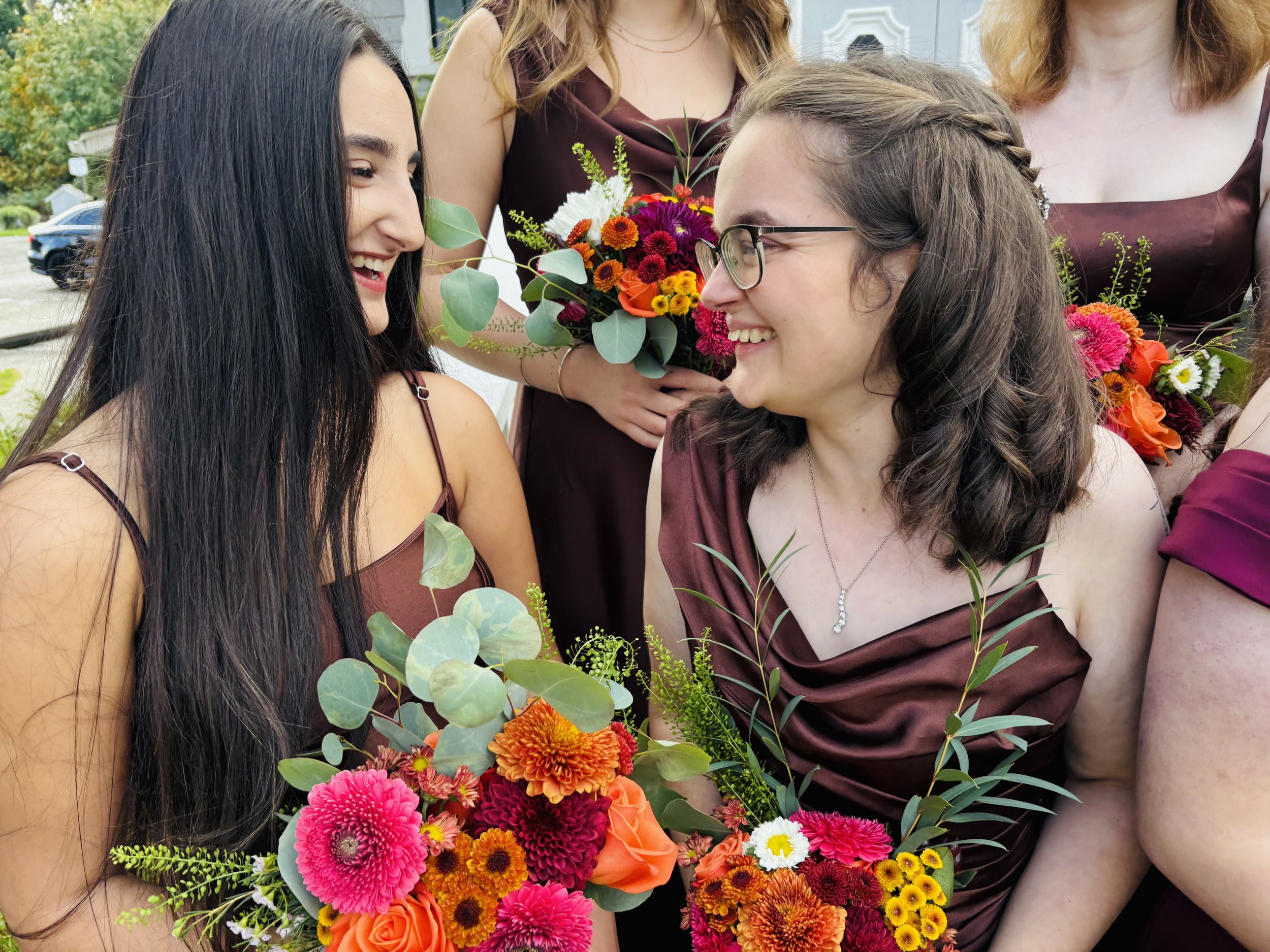 A group of women in burgundy dresses holding colorful bouquets of flowers, smiling and looking at each other, outdoors during daytime.