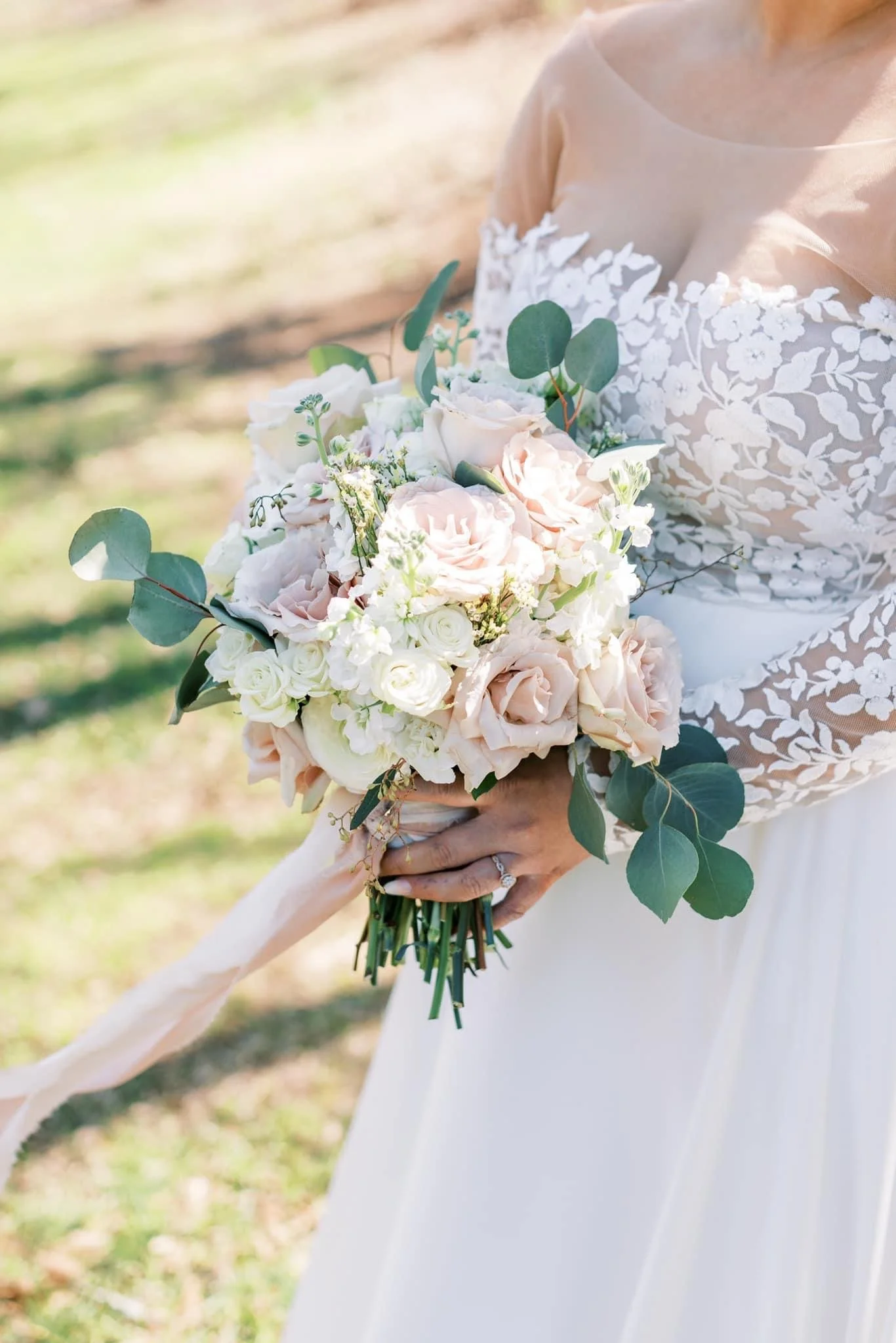 A bride holding a bouquet of blush and white roses, green eucalyptus leaves, and white flowers outdoors.
