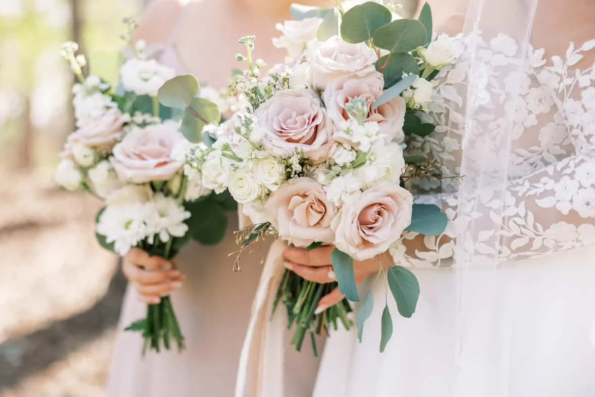 A bride holding a bouquet of pink and white roses with greenery, in front of her lace wedding dress.