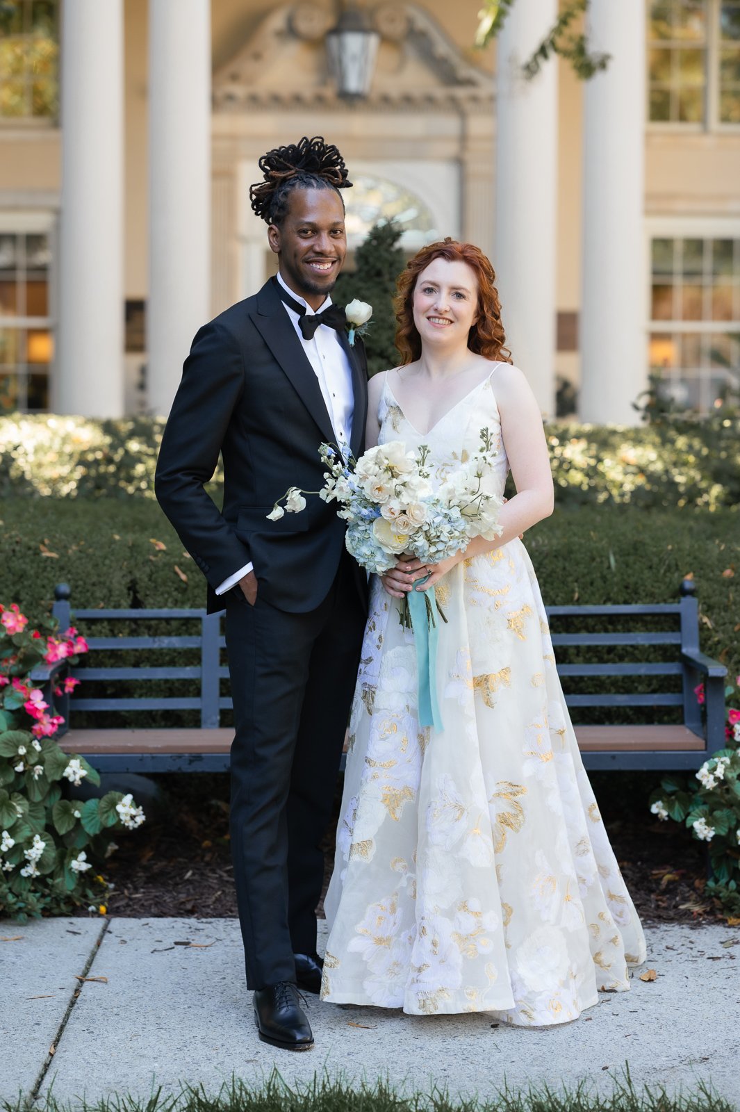 A couple in wedding attire, standing outdoors in front of a building with columns, smiling at the camera. The groom wears a black tuxedo with a bow tie, and the bride wears a white floral gown holding a bouquet of flowers.