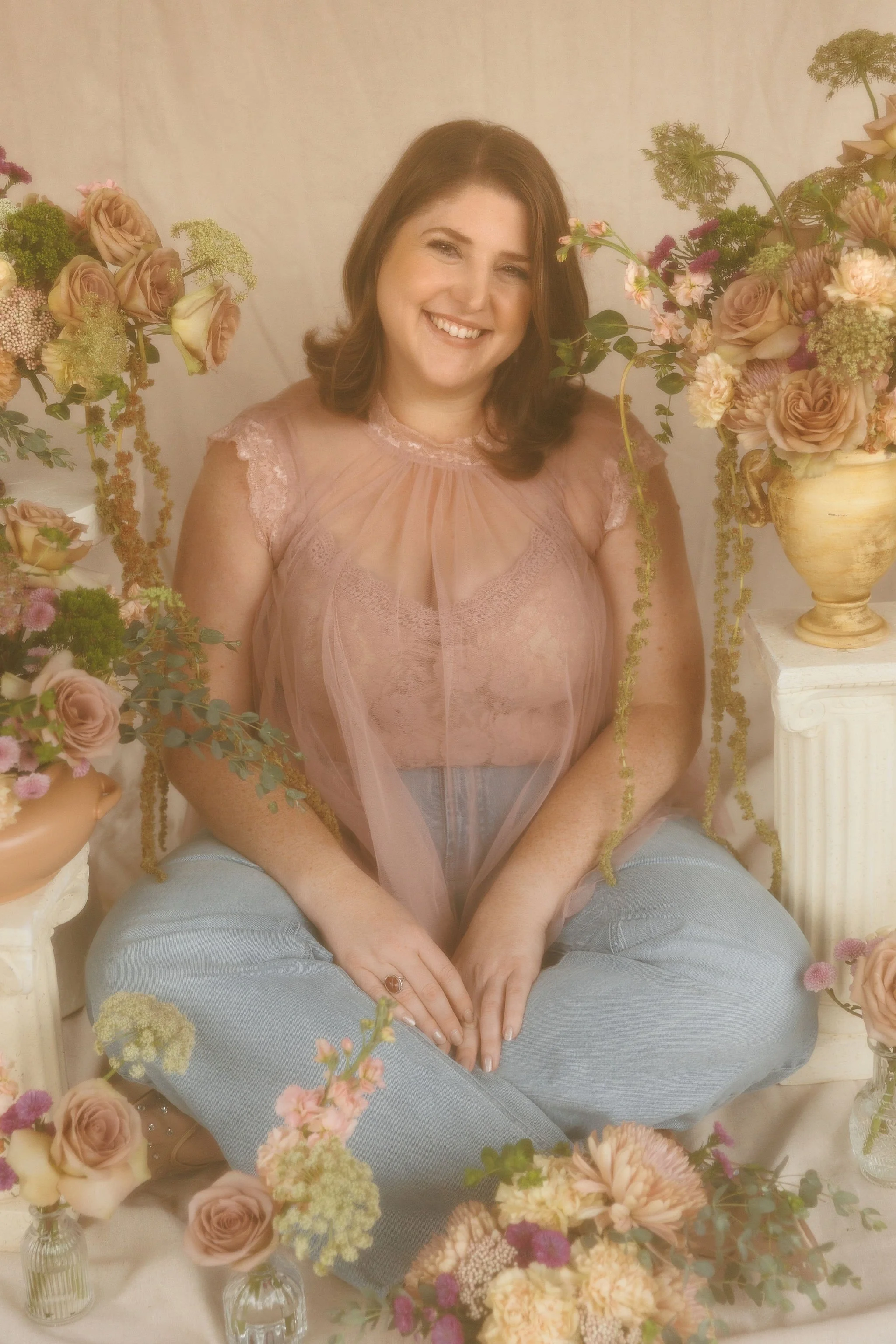 A smiling woman with brown hair, sitting cross-legged among various pastel-colored flowers in vases, with a white backdrop.