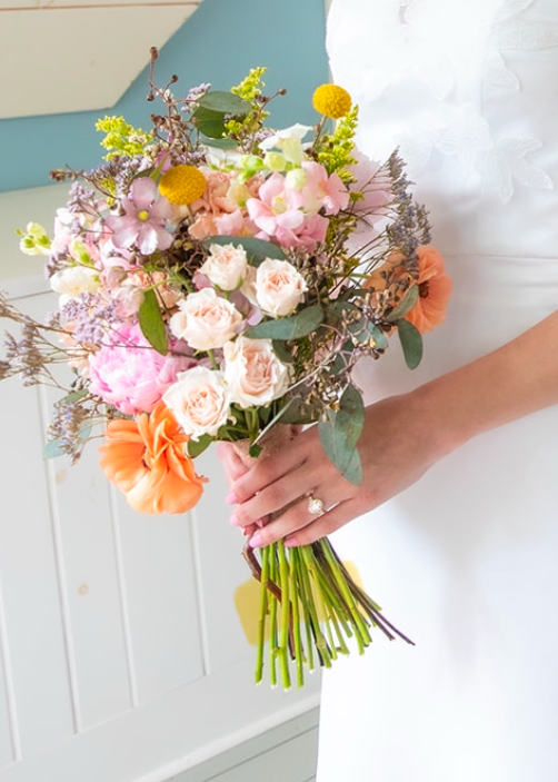 Person holding a colorful bouquet of flowers including pink roses, orange blossoms, yellow billy balls, and light purple flowers.