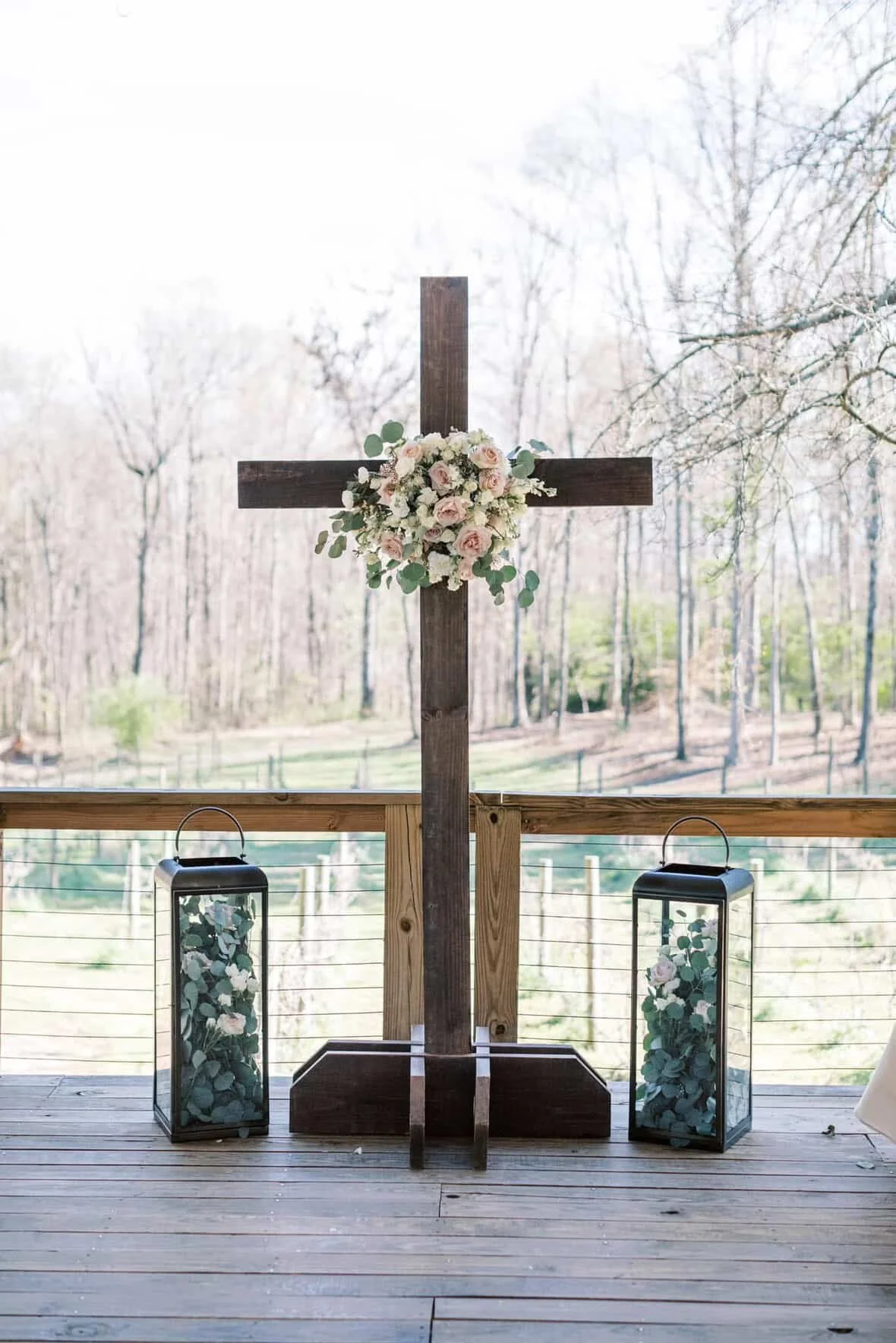 Wooden cross decorated with a bouquet of white and pink roses and greenery, flanked by two lanterns with floral arrangements inside, on a wooden deck overlooking a tree-filled landscape.