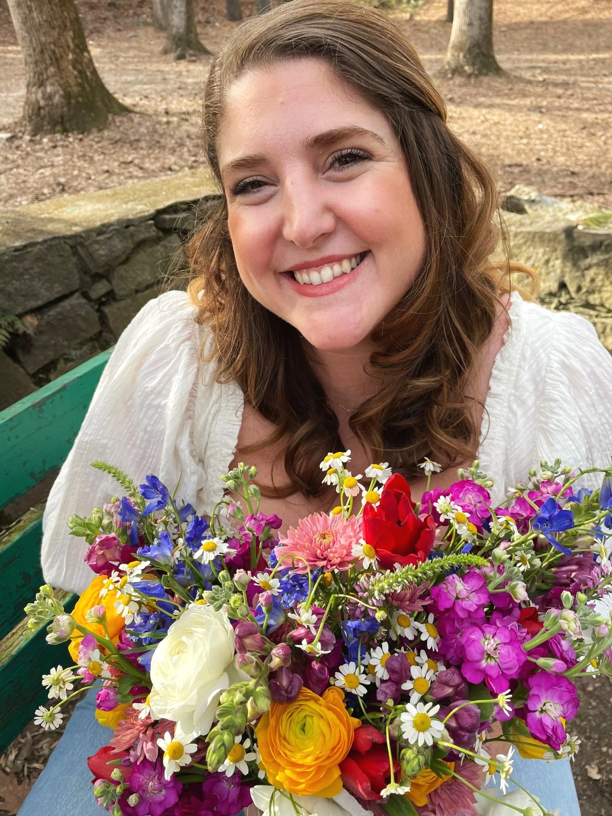 Smiling woman with brown wavy hair holding a colorful bouquet of flowers outdoors in a park.