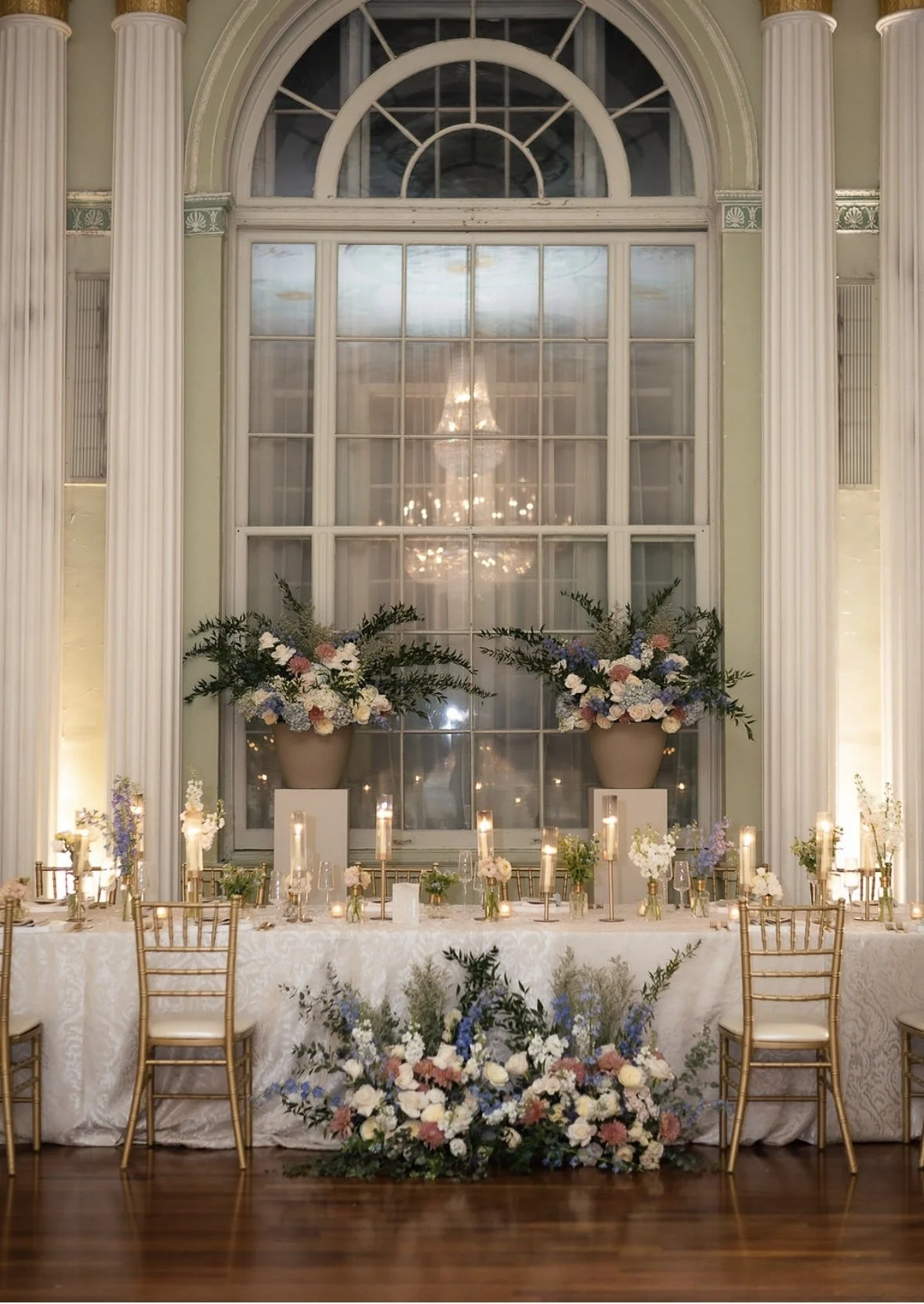 Elegant event table with floral arrangements, candles, and gold chairs in a decorated room with large window and chandelier reflection.