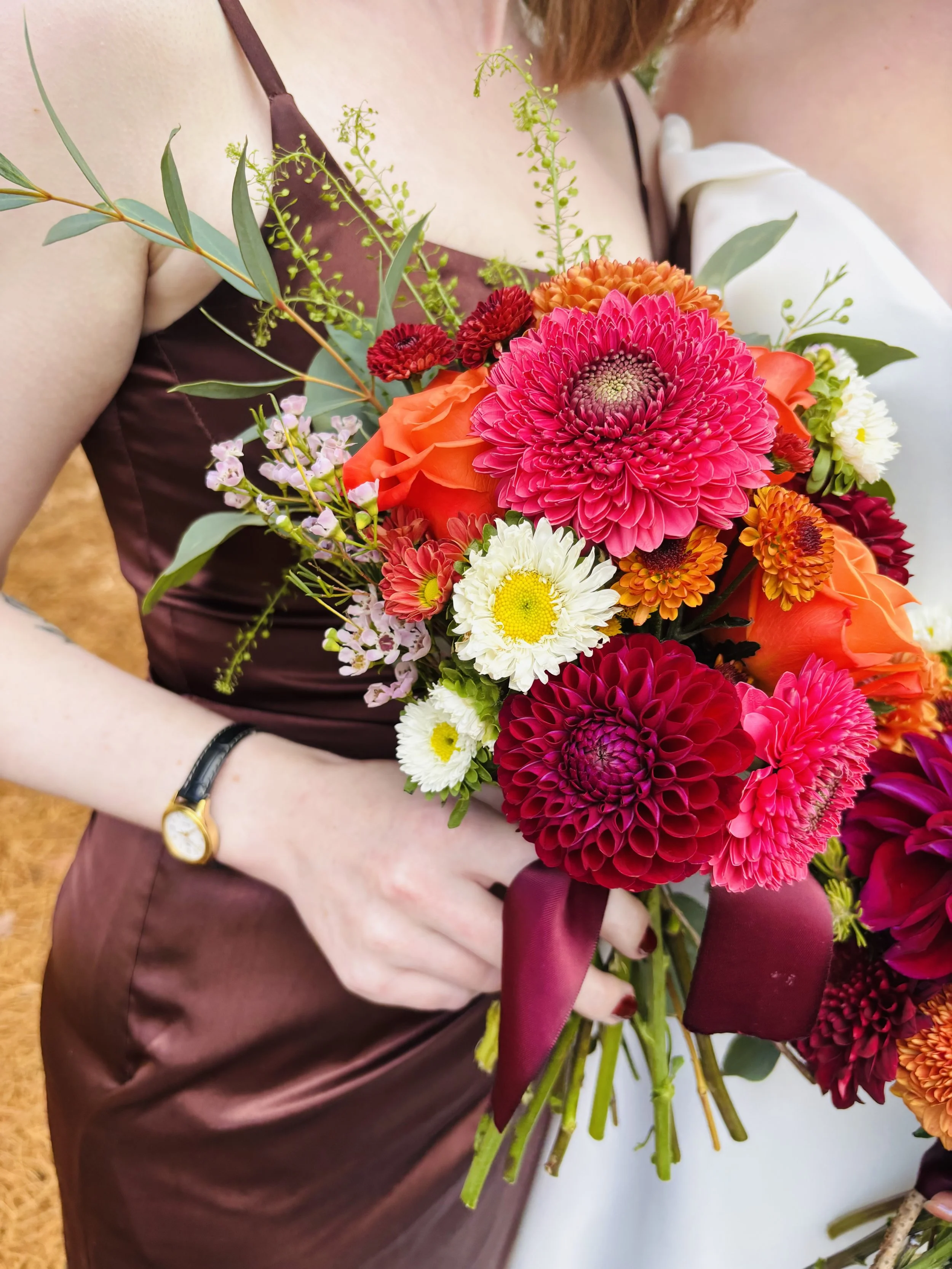 A woman holding a colorful bouquet of flowers, including pink and red dahlias, white daisies, orange roses, and greenery, wearing a brown satin dress.