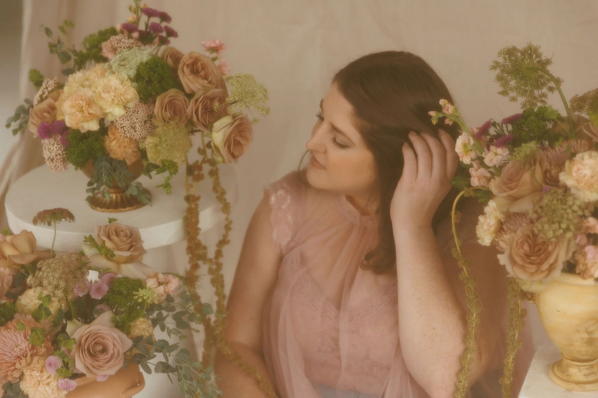 A woman with brown hair wearing a pink lace dress, surrounded by floral arrangements including roses, hydrangeas, and greenery in vases, as she touches her hair and looks down.