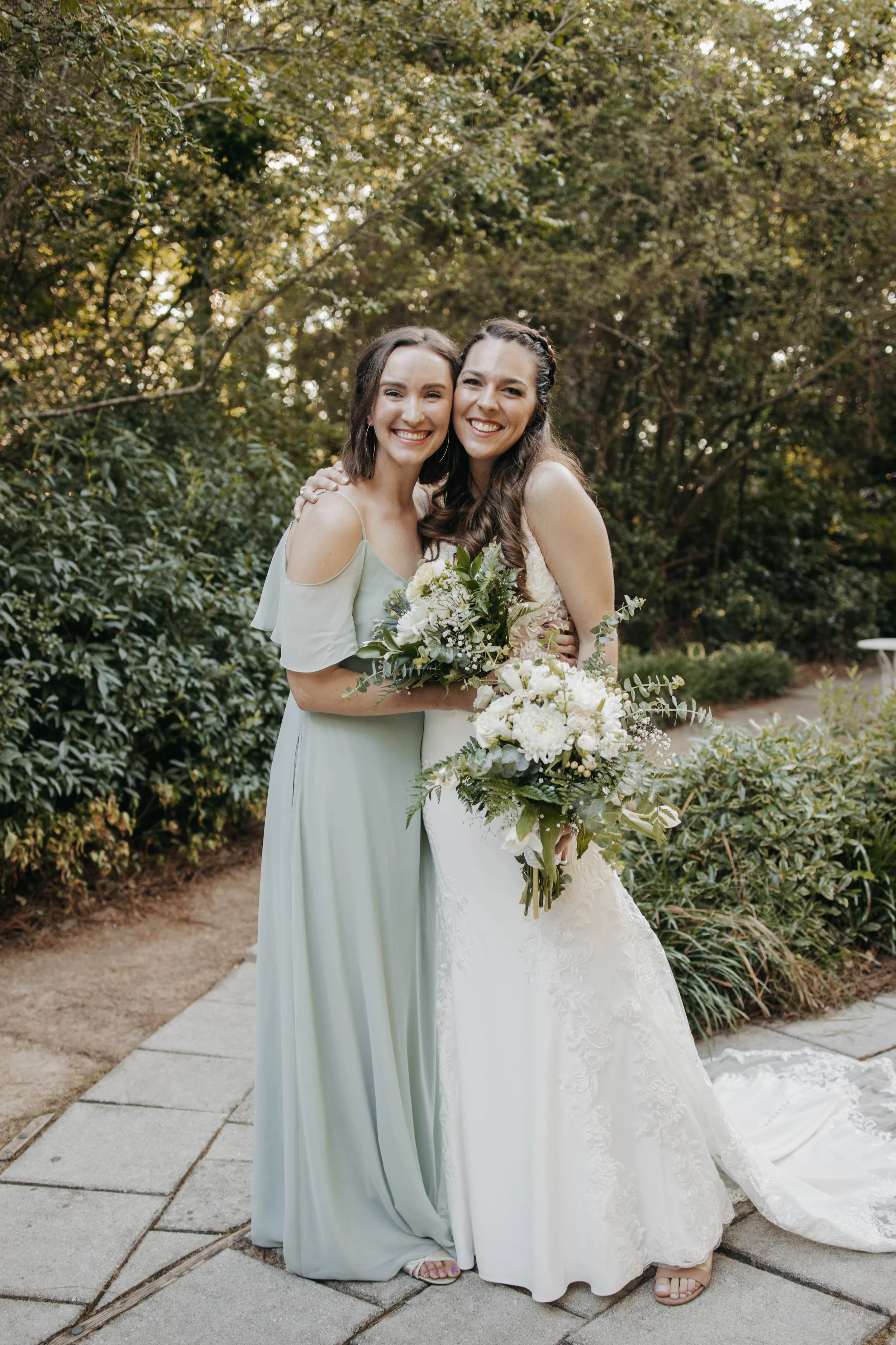 Two women in wedding attire smiling and holding bouquets, standing outdoors on a stone path surrounded by greenery.