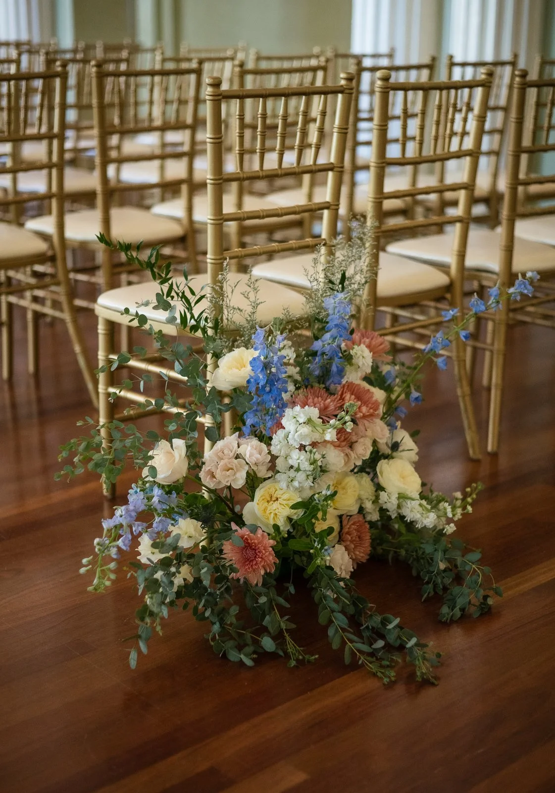 A floral arrangement with white, pink, blue, and yellow flowers, placed in front of gold chiavari chairs on a wooden floor, likely set up for a wedding or special event.