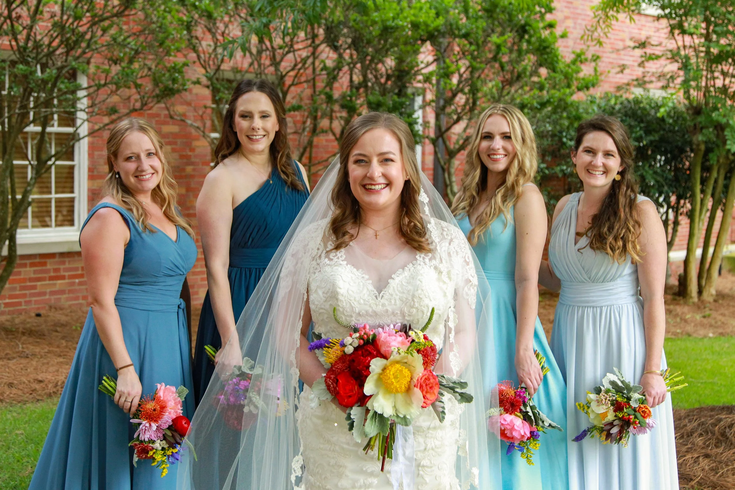 Bride in a white lace wedding dress holding a bouquet, surrounded by five bridesmaids in blue dresses holding smaller bouquets, outdoors in front of a brick building with trees.