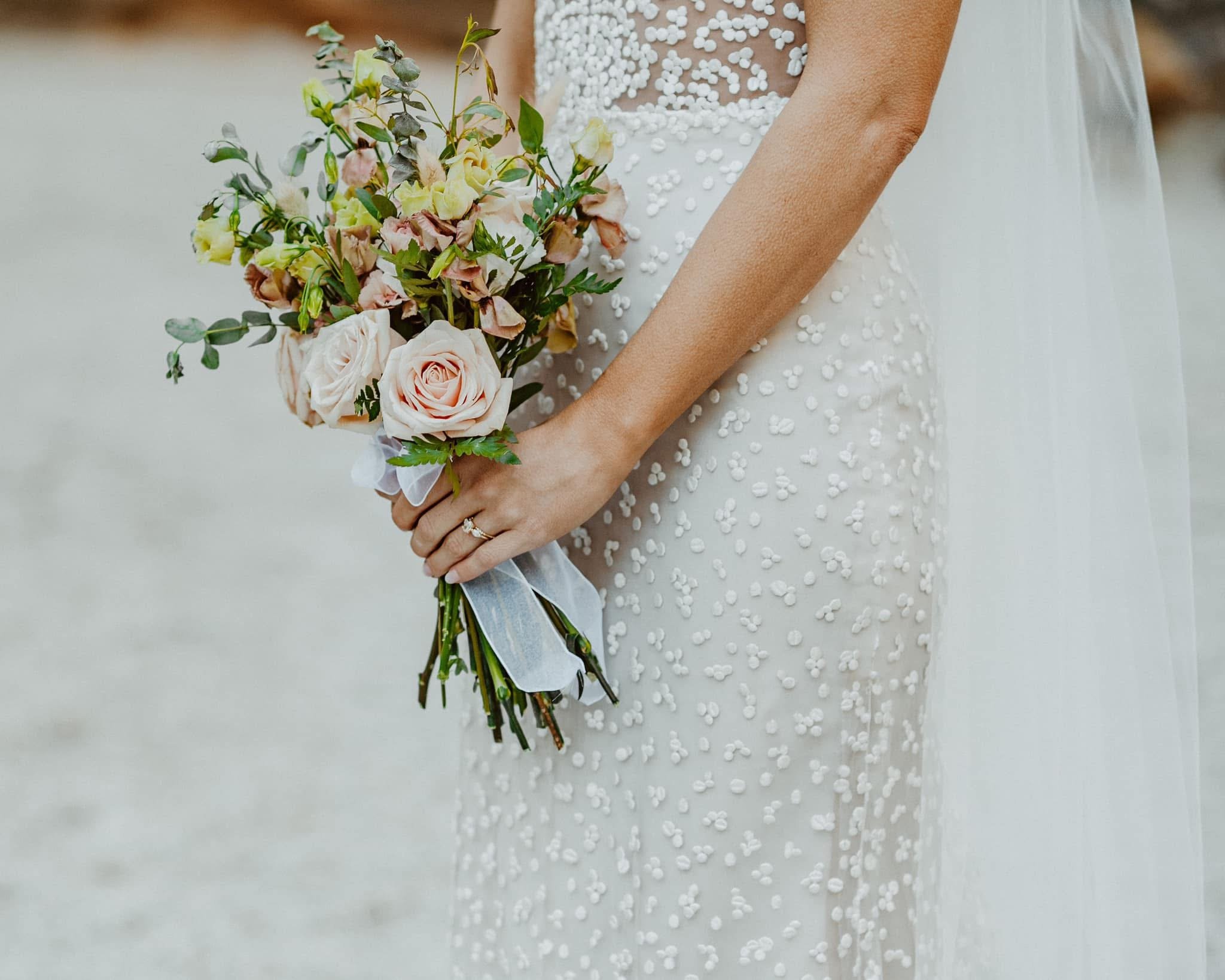 A bride holding a bouquet of light pink roses and green foliage, wearing a white wedding dress with 3D floral appliqués and a sheer top with pearl embellishments.