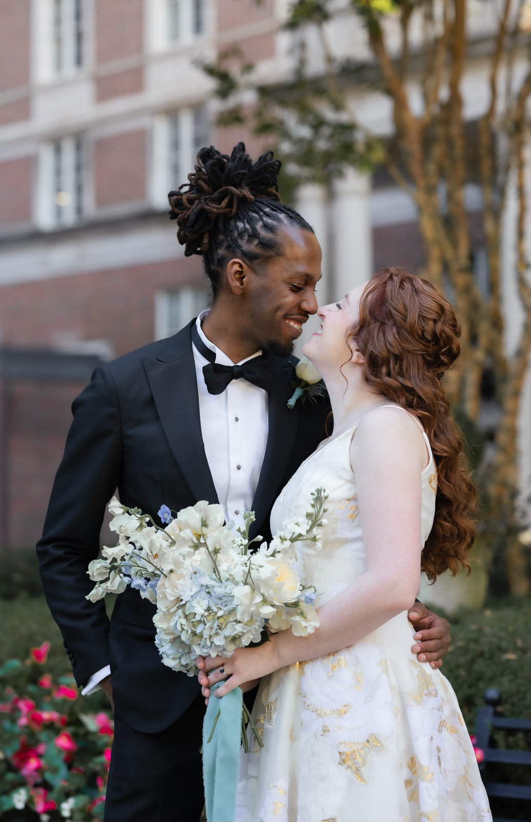 A couple on their wedding day, with the groom wearing a black tuxedo and the bride in a white dress holding a bouquet, standing close together and smiling outdoors.