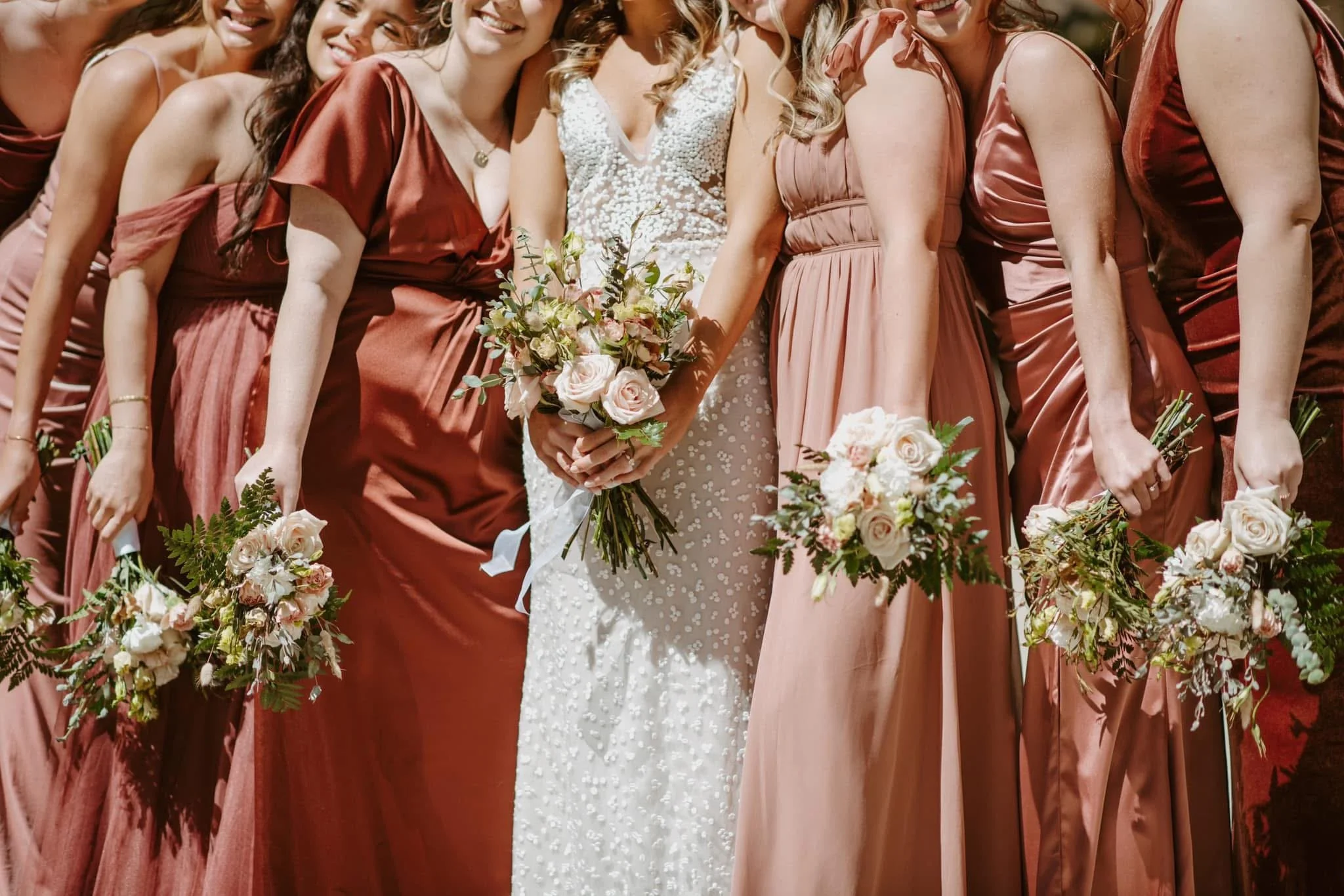 Group of women in bridesmaid dresses and a bride in a white wedding gown holding flowers, standing close together at a wedding event.