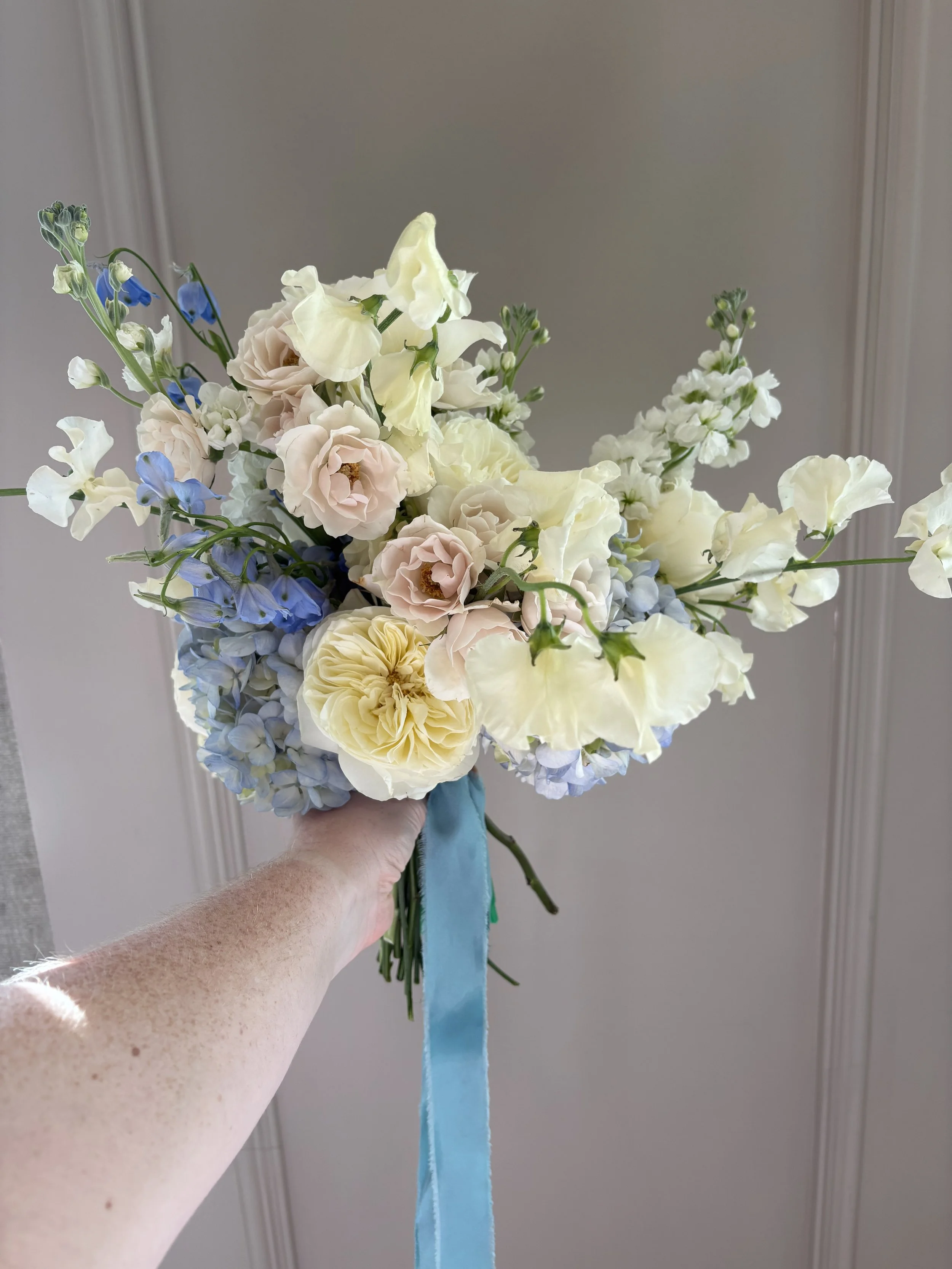 Hand holding a bouquet of white, cream, and light pink flowers with blue accents and a blue ribbon, against a plain gray wall background.