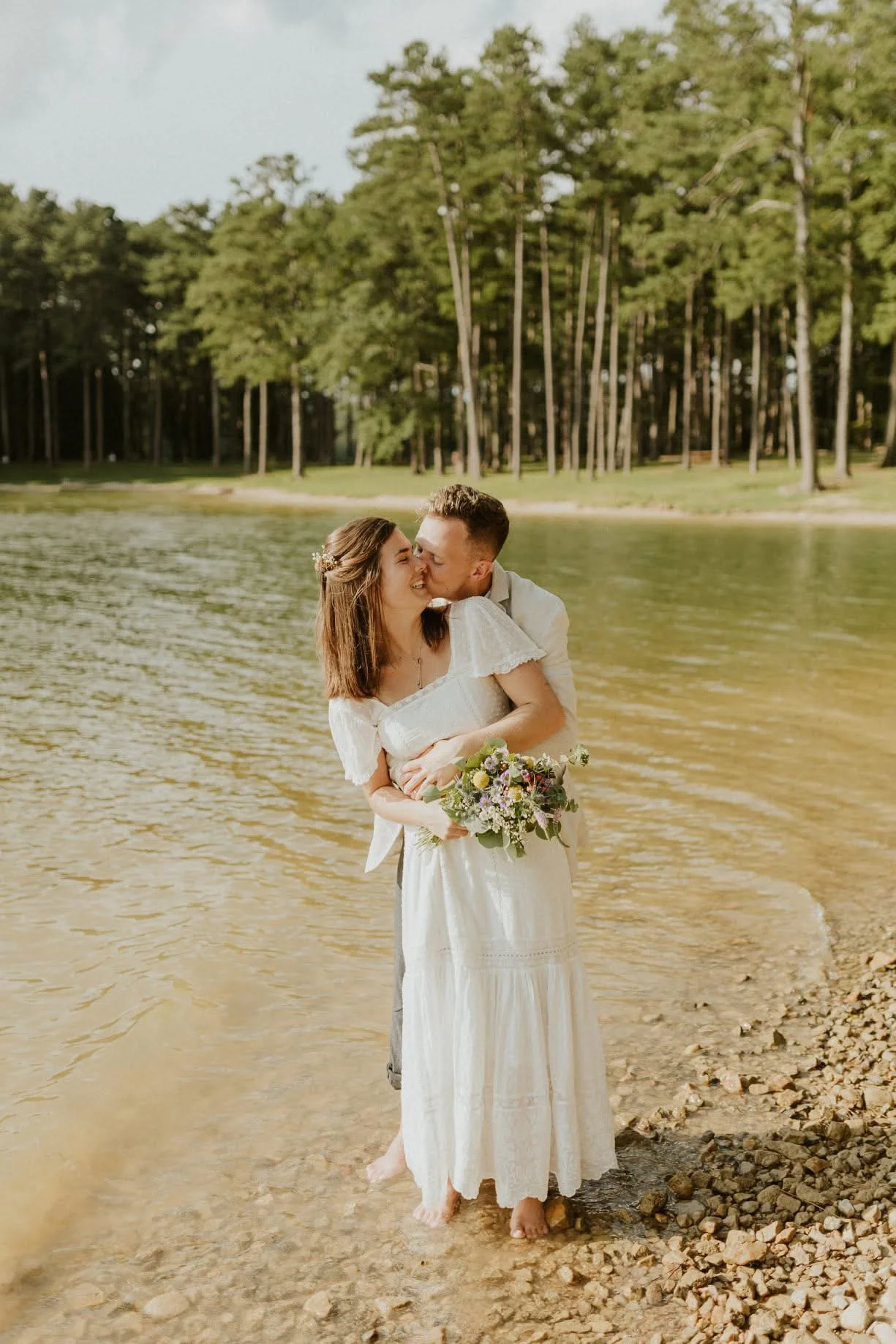 A newlywed couple sharing a kiss by a lakeside, with the bride holding a bouquet of flowers and both standing barefoot on pebbles.