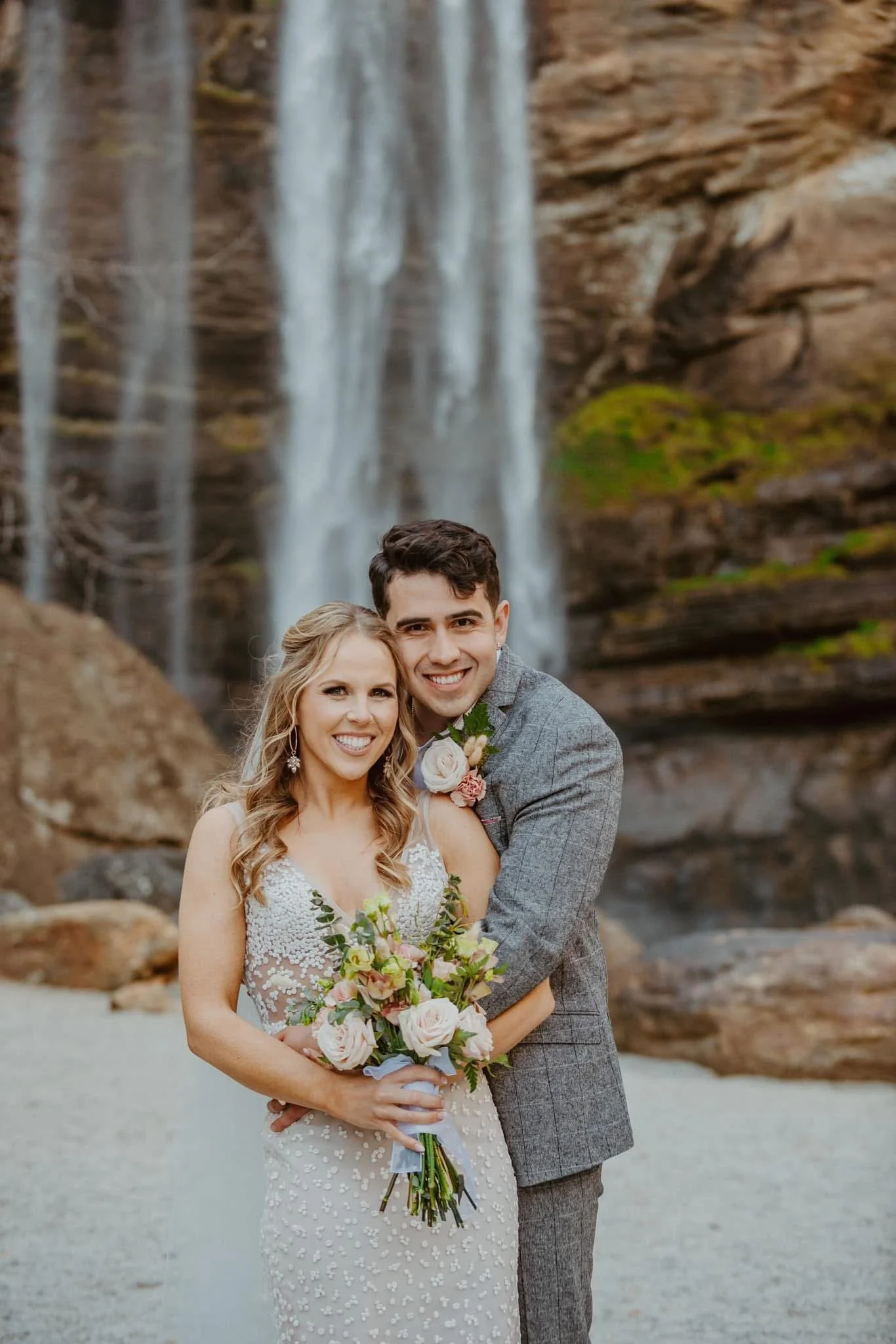 A smiling bride and groom pose in front of a waterfall, with the bride holding a bouquet of pale pink roses and greenery, and the groom in a gray suit.
