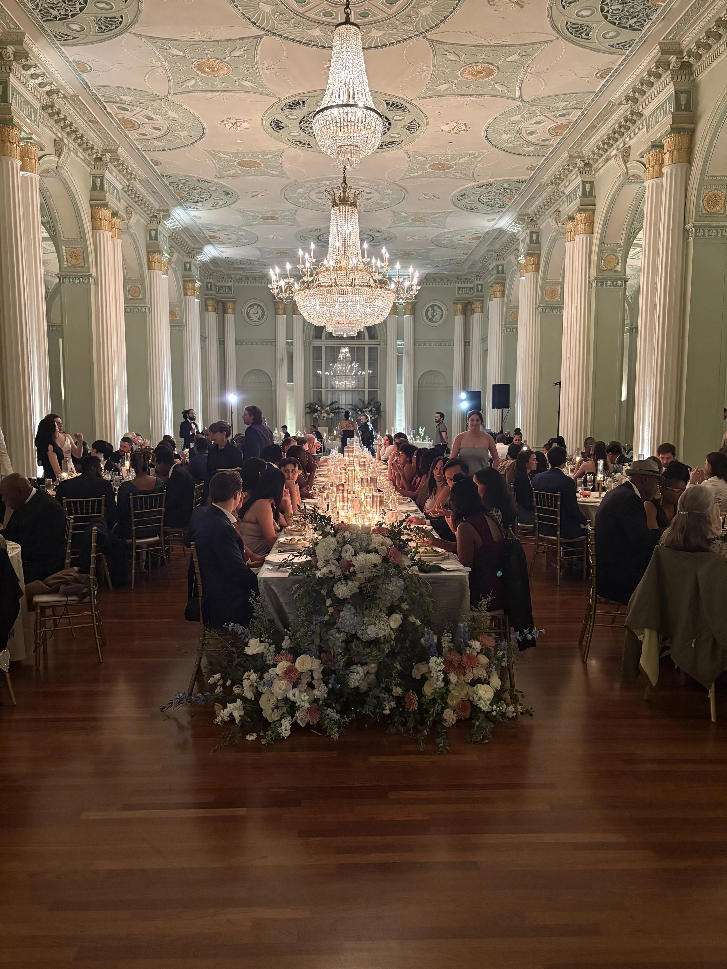 An elegant banquet hall with a long dining table decorated with floral arrangements, chandeliers hanging from the ornate ceiling, and guests seated on either side of the table at a formal event.