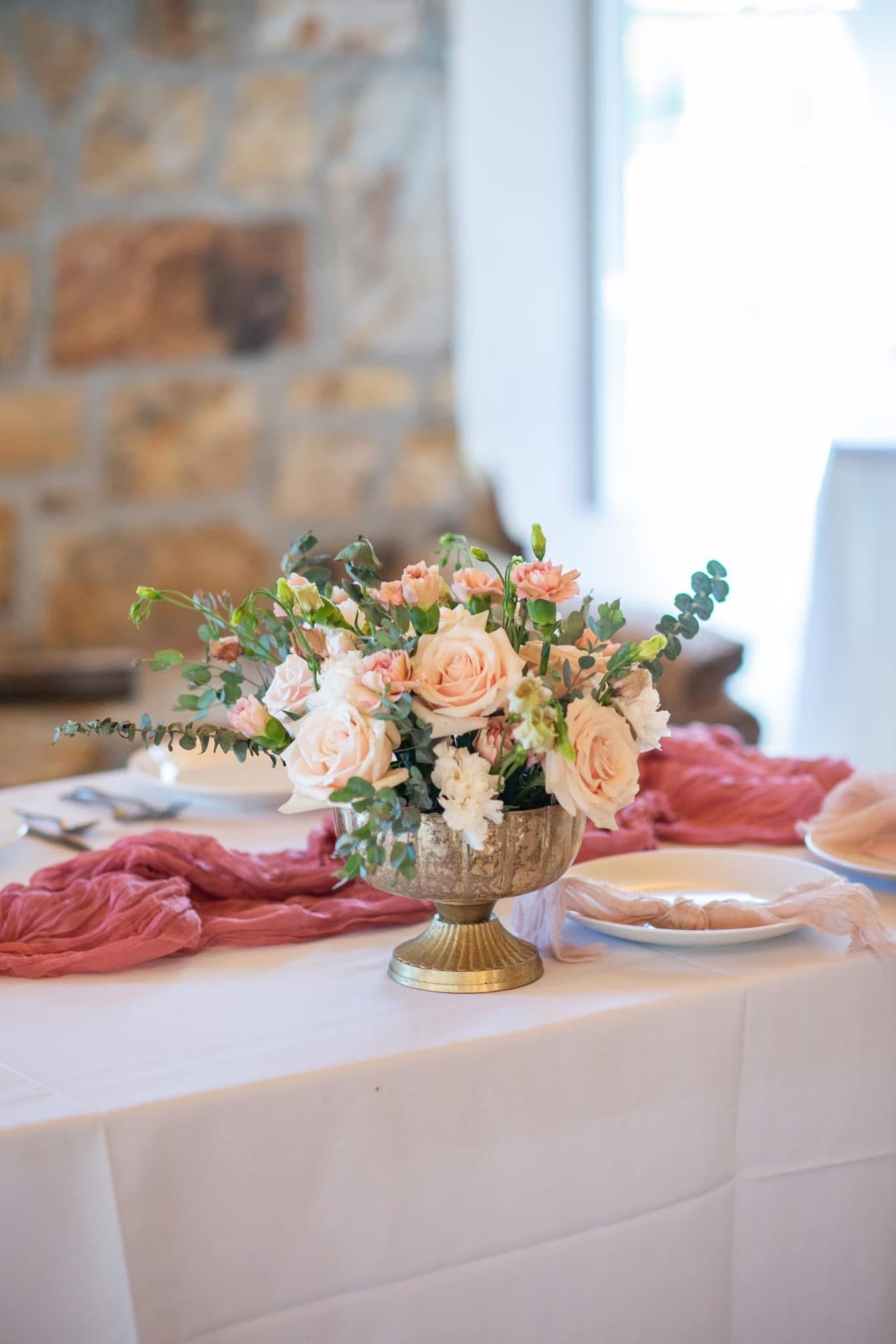 Elegant table setting with a floral centerpiece featuring pink roses and greenery, soft pink table runner, white plates, and silverware, with a rustic stone wall in the background.