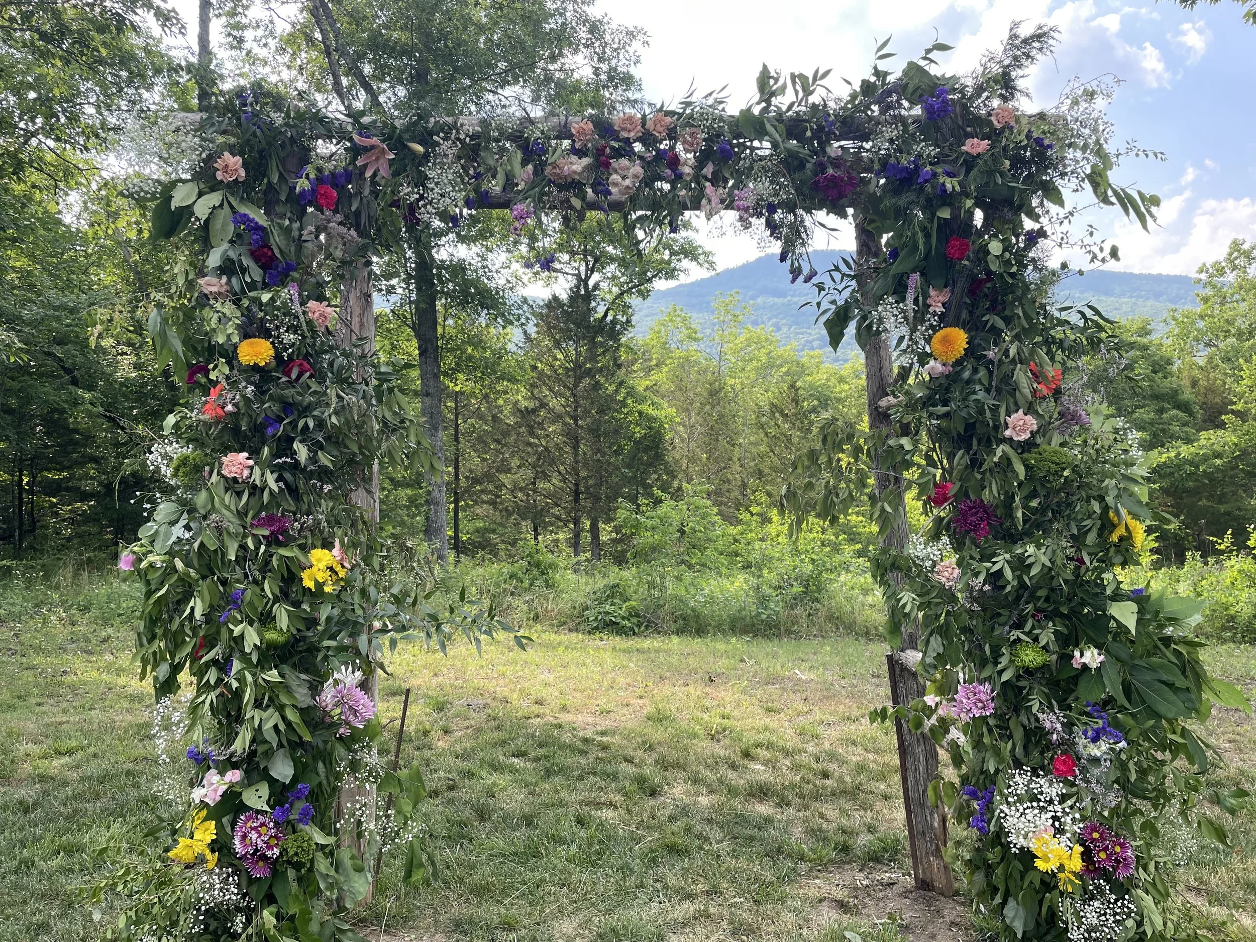 A wooden arch decorated with multicolored flowers and greenery, set outdoors in a grassy area with trees and mountains in the background.