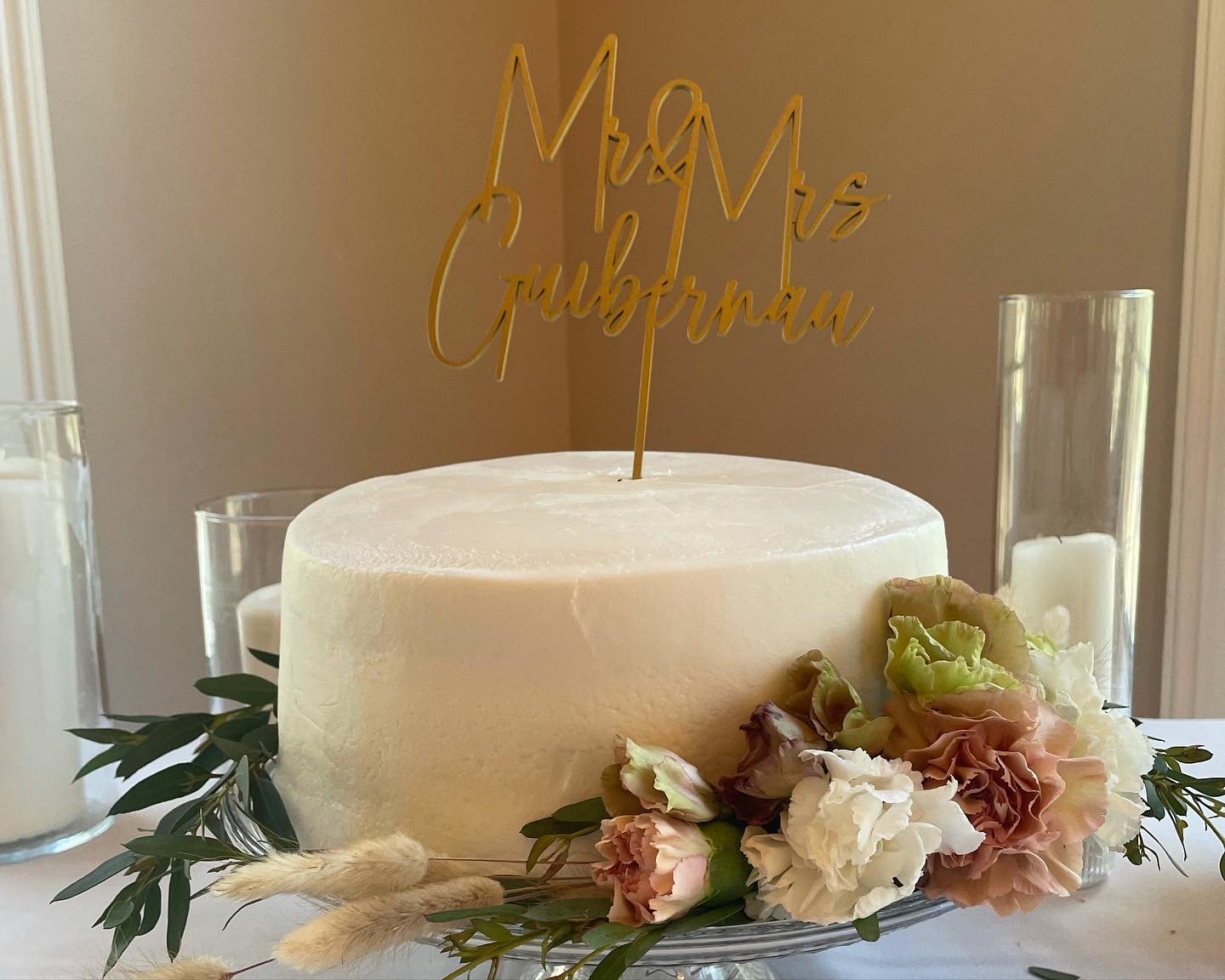 A white wedding cake with a gold topper reading 'Mr & Mrs Guerrino', decorated with flowers and greenery, on a glass plate surrounded by candles.