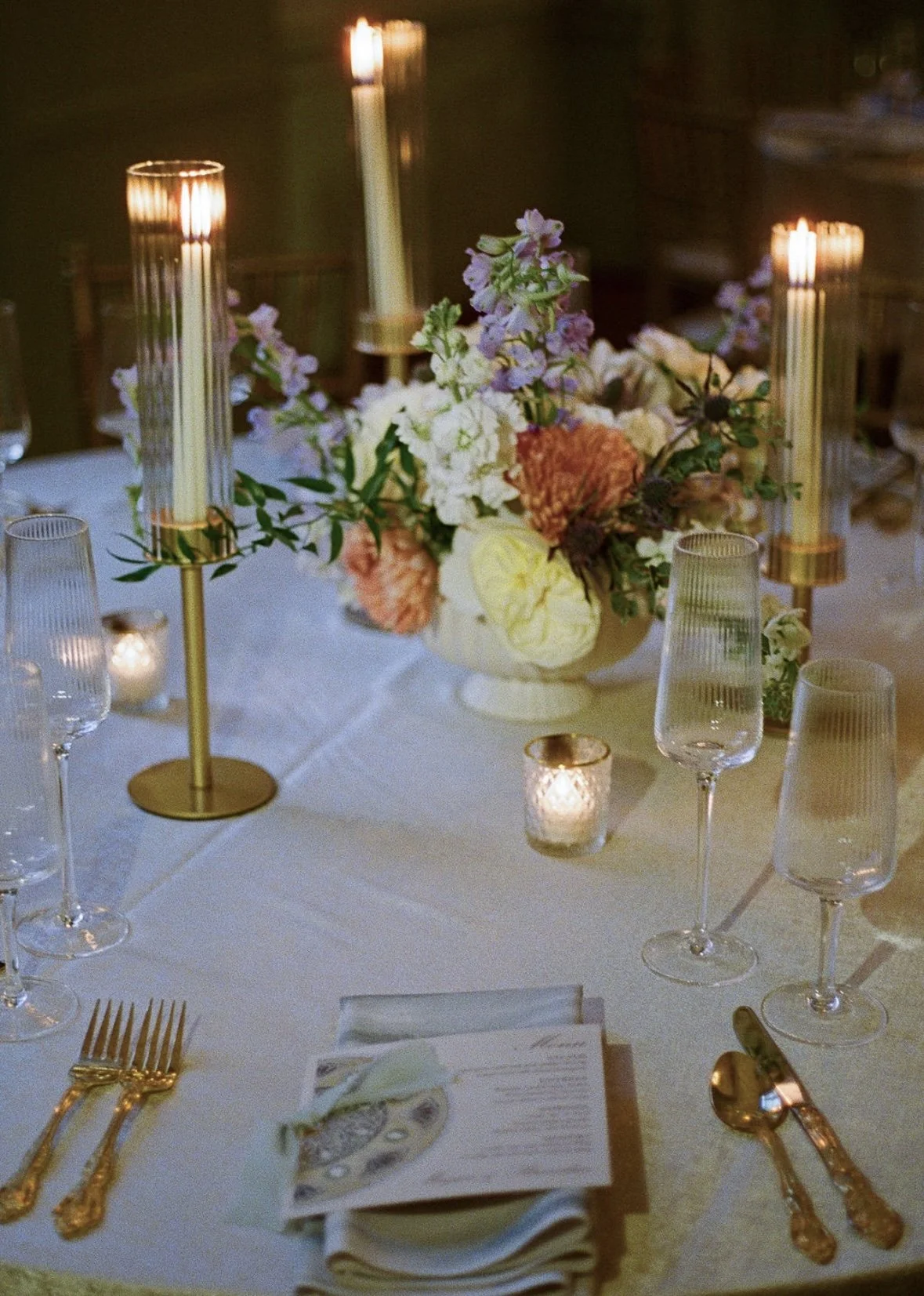 Elegant table setting with gold utensils and candlelight, featuring a floral centerpiece with pink, white, and purple flowers on a white tablecloth.
