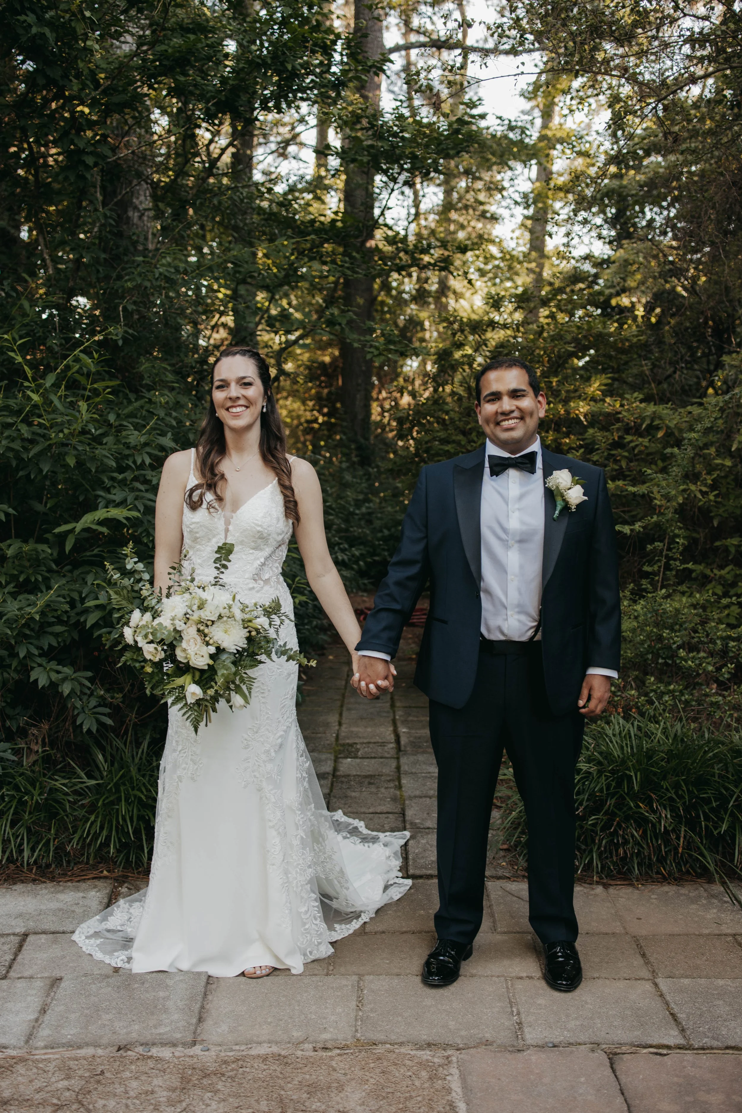 A newlywed couple holding hands outdoors on a stone pathway surrounded by greenery, smiling at the camera. The bride wears a white lace wedding dress and holds a bouquet, the groom wears a dark suit with a bow tie and a white boutonniere.
