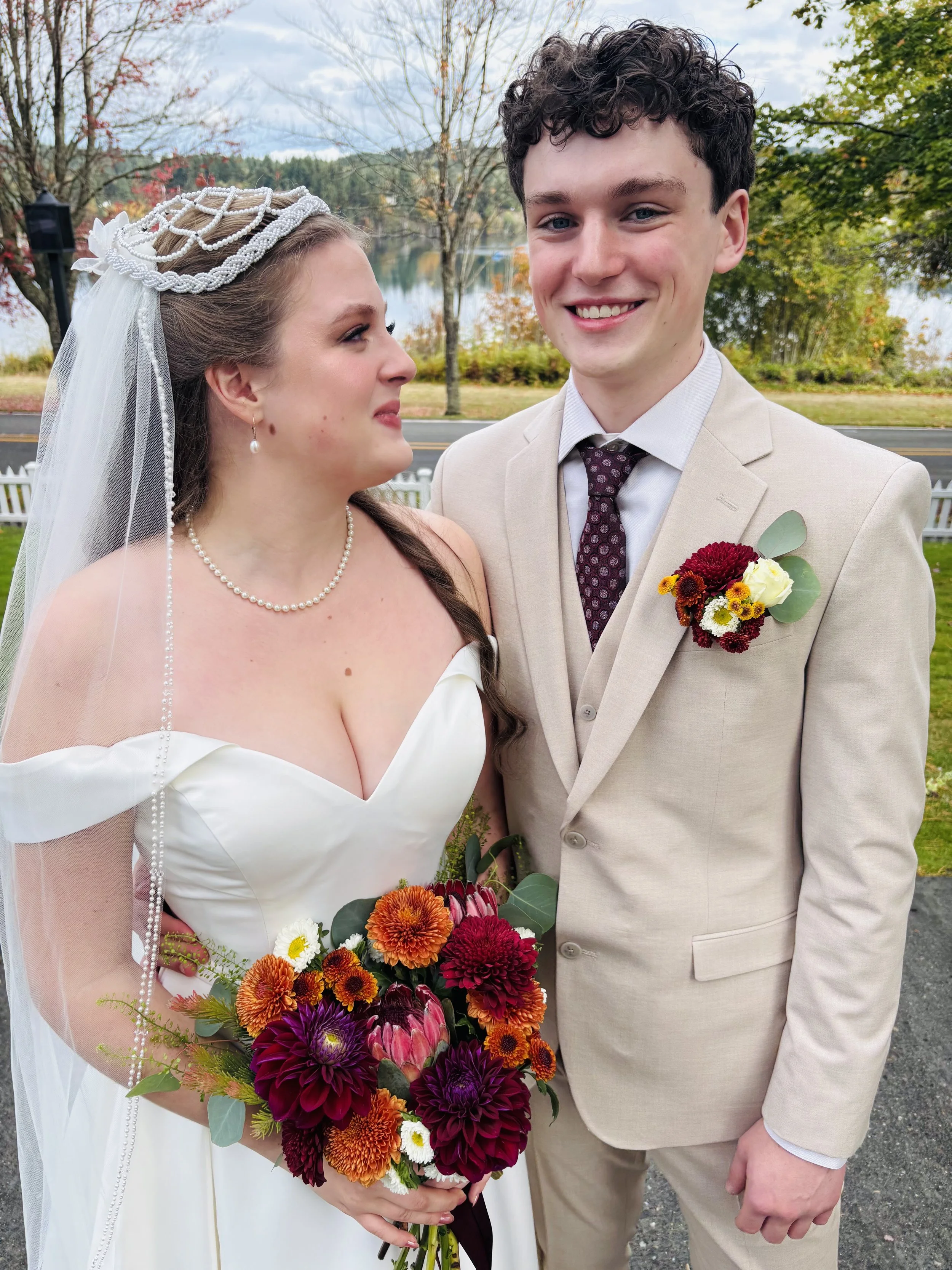 Bride and groom standing outdoors on a rainy day, smiling and looking at each other. The bride is holding a colorful bouquet of flowers, wearing a white wedding dress and veil with pearl accessories. The groom is wearing a beige suit with a dark, pat