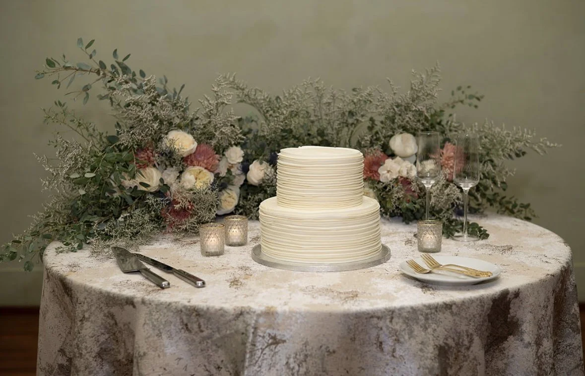 Wedding cake on table with floral arrangement, candles, glasses, and cutlery.