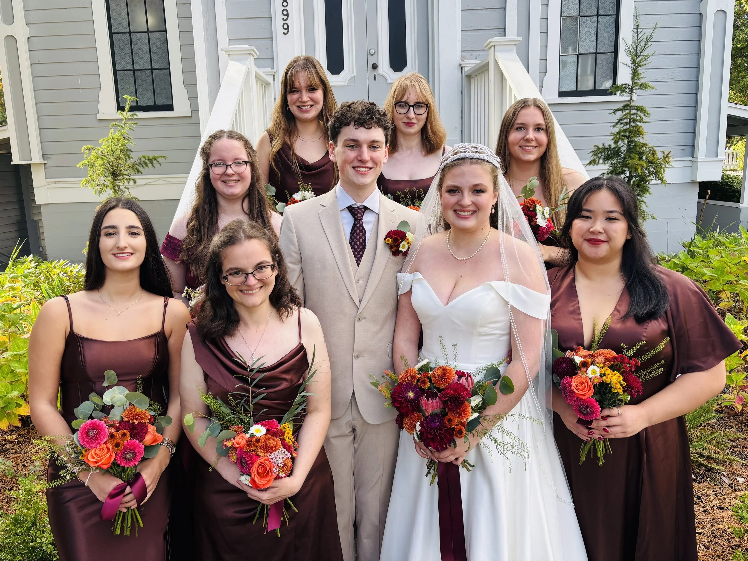 A wedding group photo with the bride, groom, and eight bridesmaids outside a house. The bride is in a white gown holding a bouquet, and the groom is in a tan suit. The bridesmaids are in matching dark burgundy dresses, each holding a bouquet. The gro