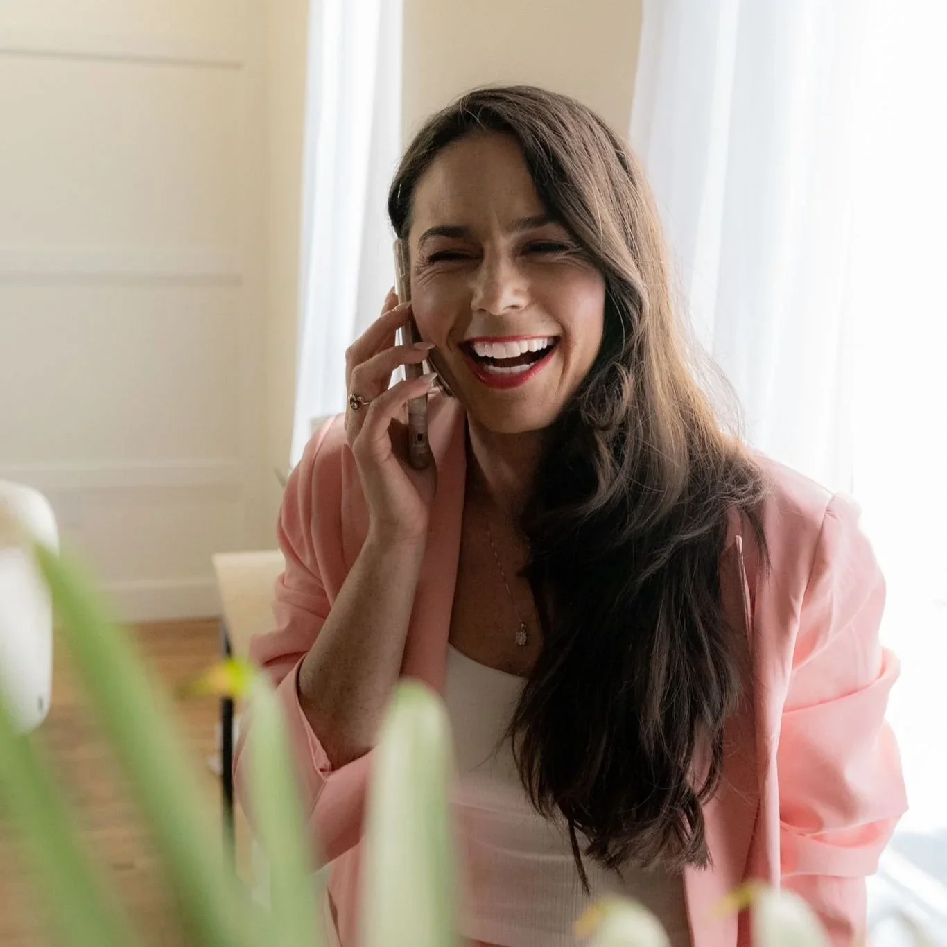 A woman with long dark hair, wearing a pink blazer and white top, is laughing while talking on the phone indoors near a window with white curtains.