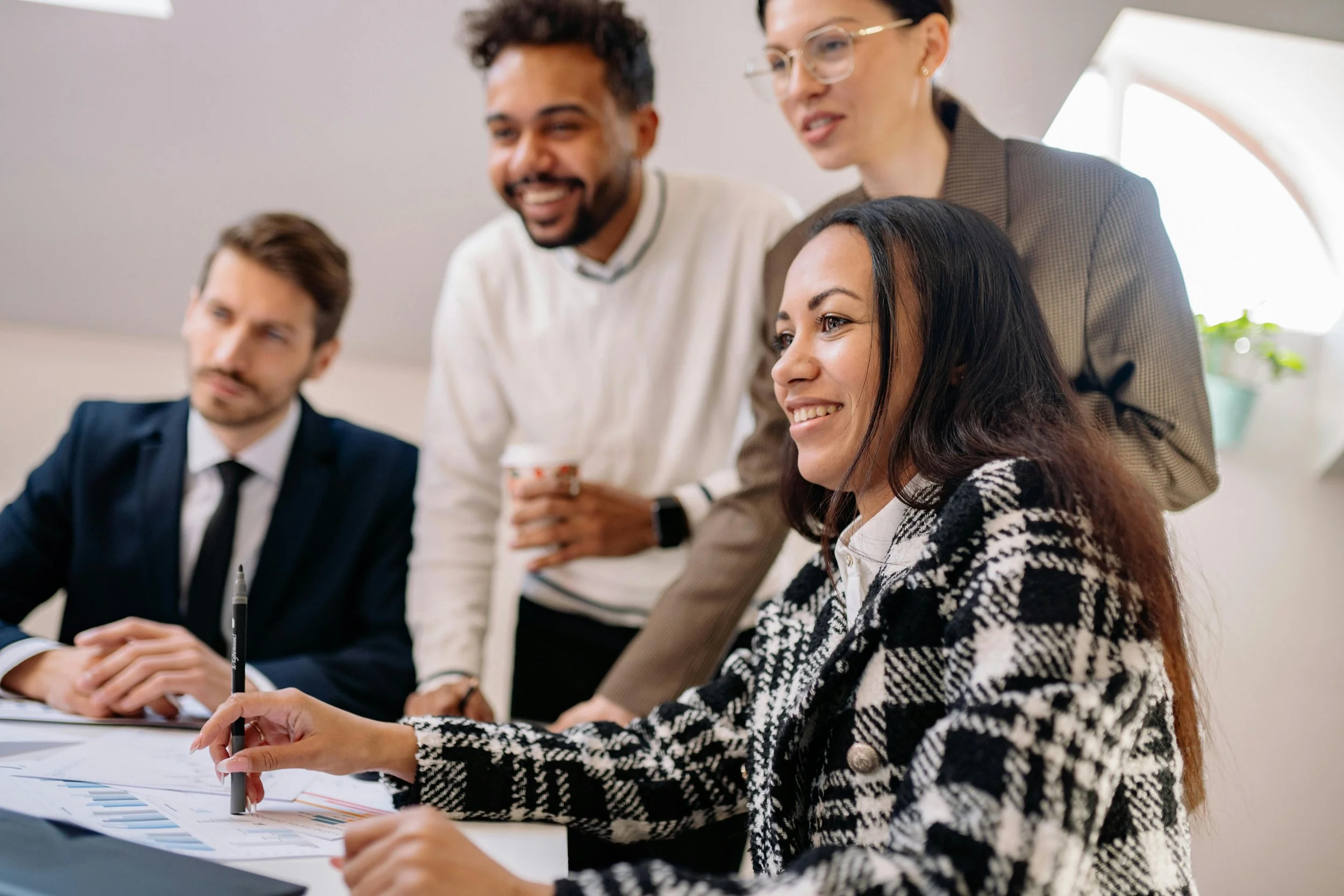 Four diverse people in a business meeting; a woman in a black and white blazer is smiling and pointing at documents, a man in a suit is sitting nearby, and two others are standing behind her, smiling and looking at the documents.