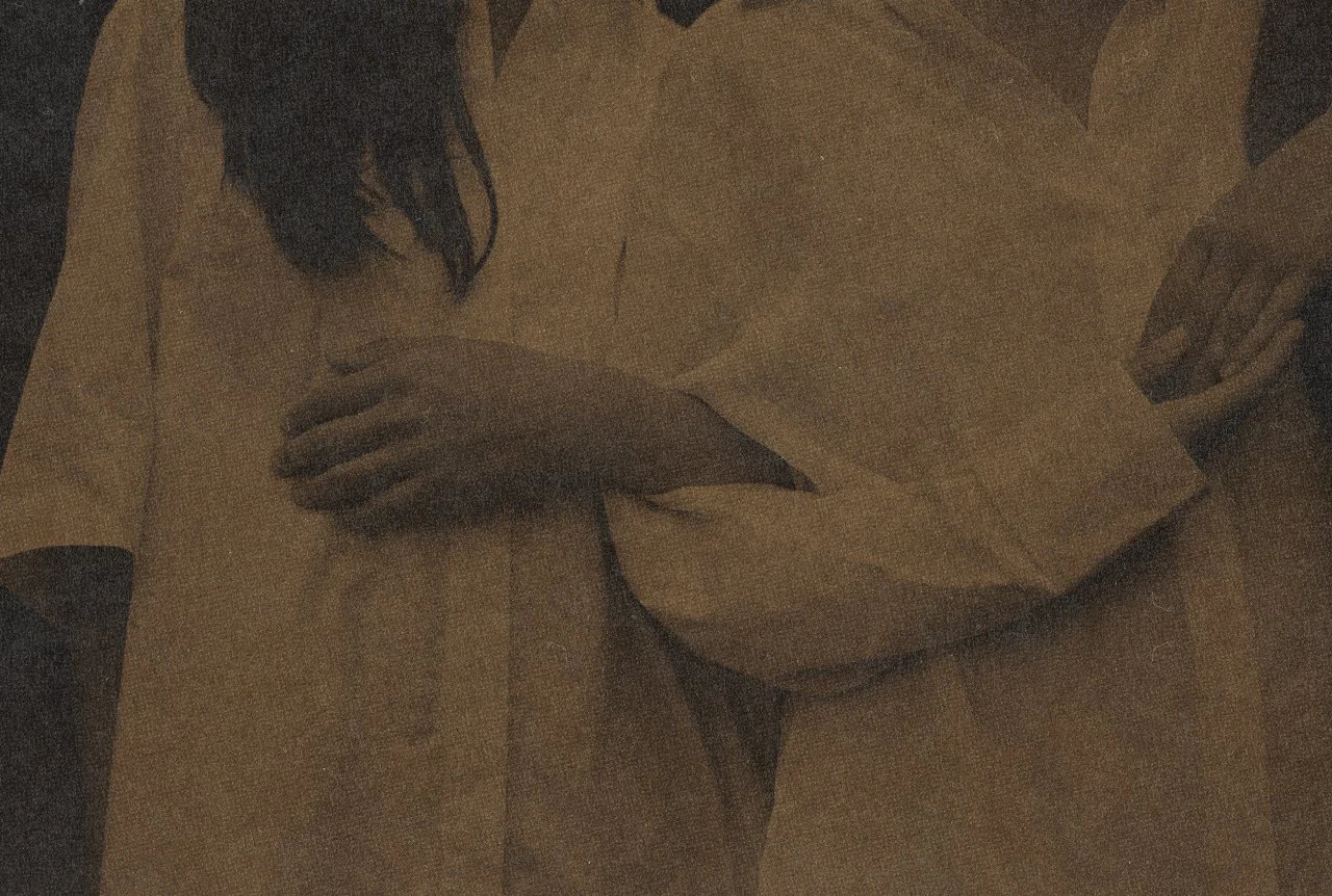 A person with long dark hair wearing a beige or light-colored shirt, with their hands resting on their chest and abdomen, standing in front of a wooden background.