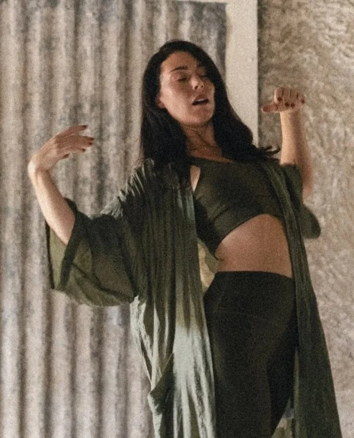 A woman with black hair, wearing a black crop top and black pants, is standing with her arms raised and eyes closed in front of a textured wall.