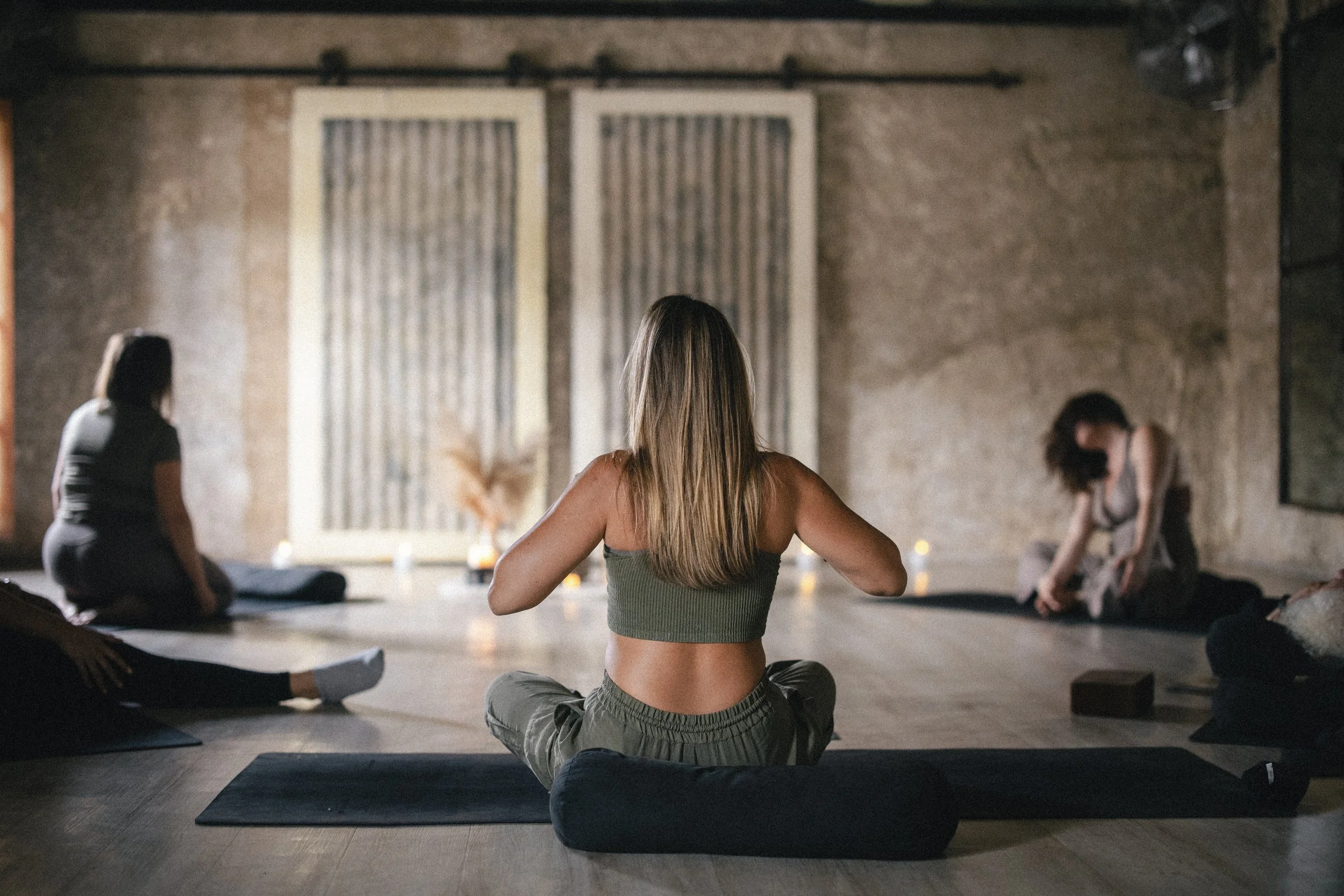 A group of women practicing yoga in a dimly lit studio with exposed brick walls and candles, sitting on yoga mats in a cross-legged position.