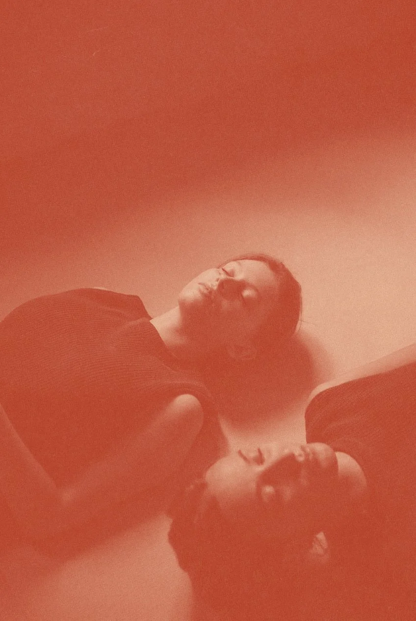 Two women lying on the floor, looking up at the camera, with a red filter over the photo.