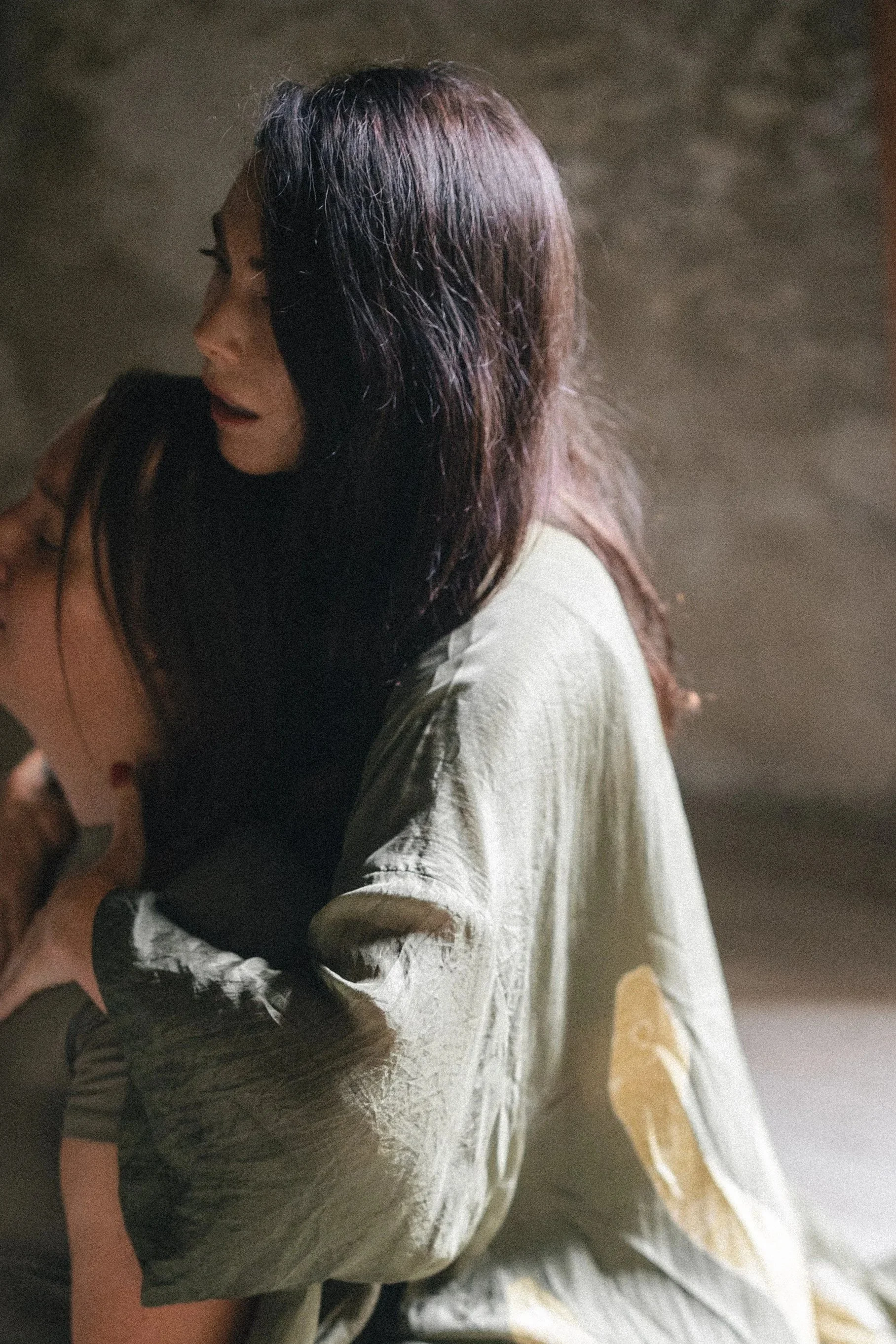 Two women hugging each other, one woman with black hair and a light-colored jacket, the other with long brown hair, in a dimly lit setting.