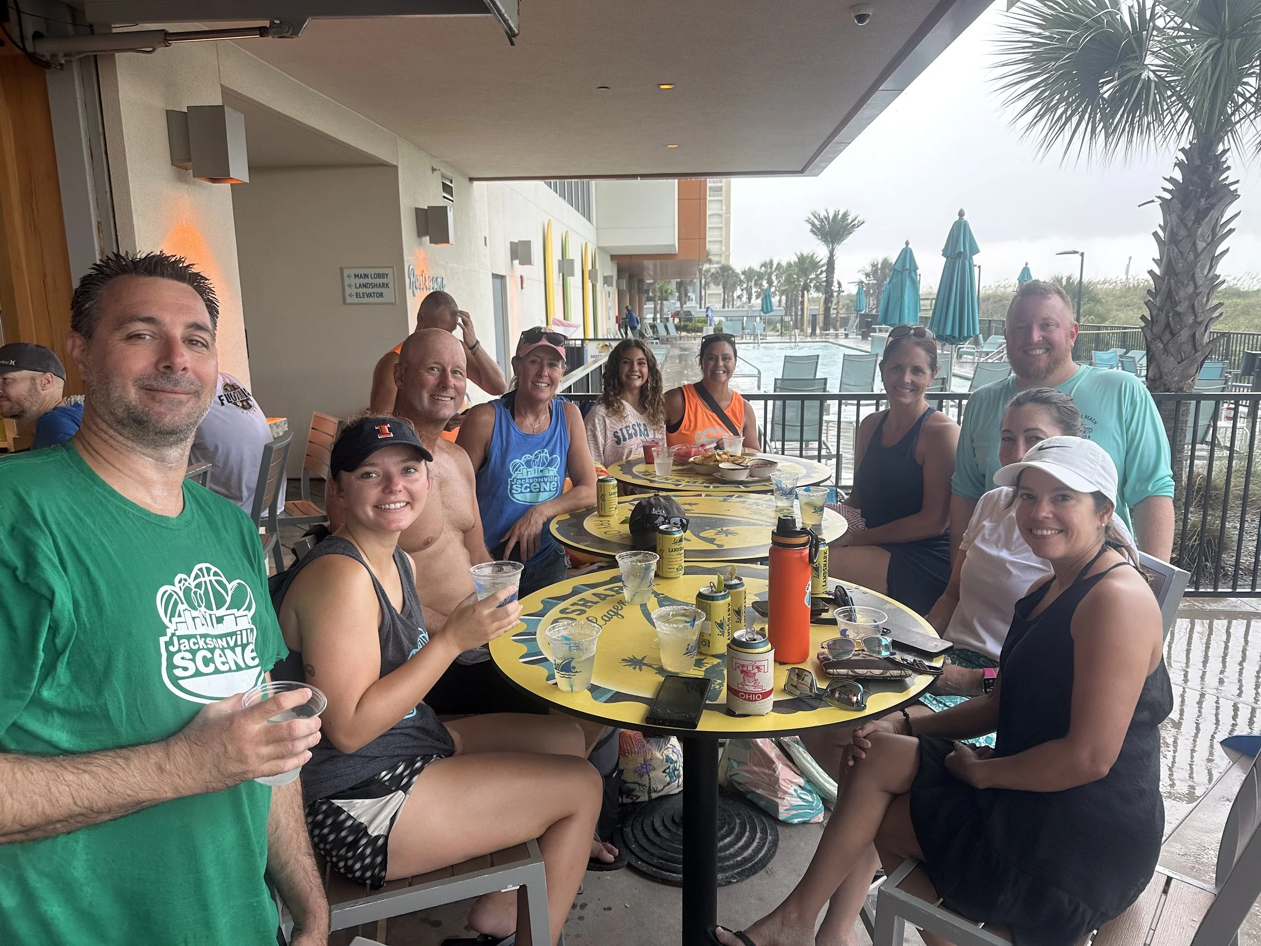 A group of eleven people gathered around a table at an outdoor restaurant near a pool area. They are smiling, holding drinks, and appear to be enjoying a casual social event. The background shows palm trees, umbrellas, and a cloudy sky.