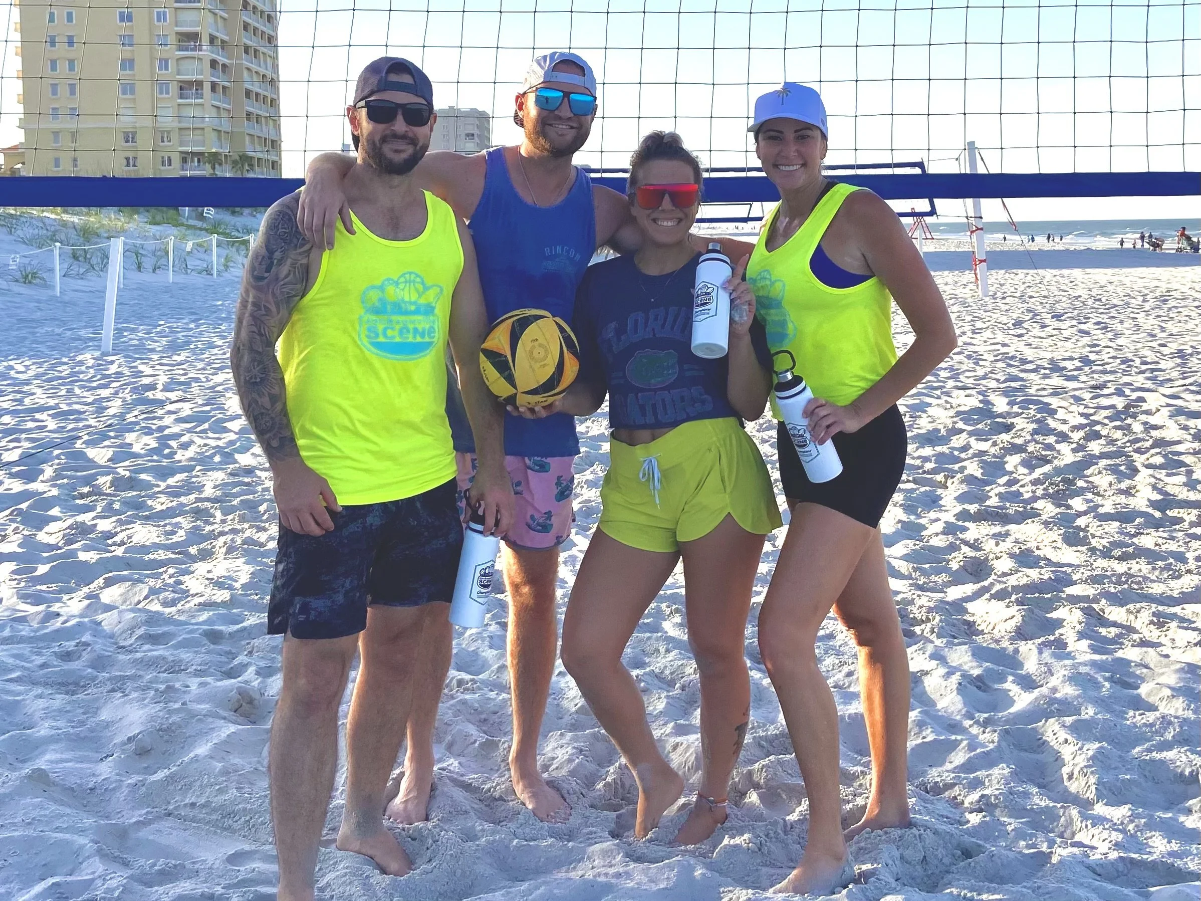 Four friends on a sandy beach standing in front of a volleyball net, smiling, holding water bottles and a volleyball, dressed in beachwear and sunglasses, with high-rise buildings and the ocean in the background.