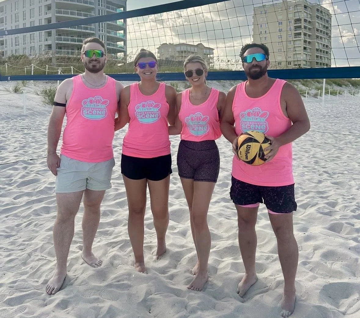 Four people standing on a sandy beach in front of a volleyball net. They are all wearing matching pink tank tops with a logo that says "Jacksonville Scene." The group consists of two men and two women, all smiling, wearing sunglasses, and holding a volleyball.