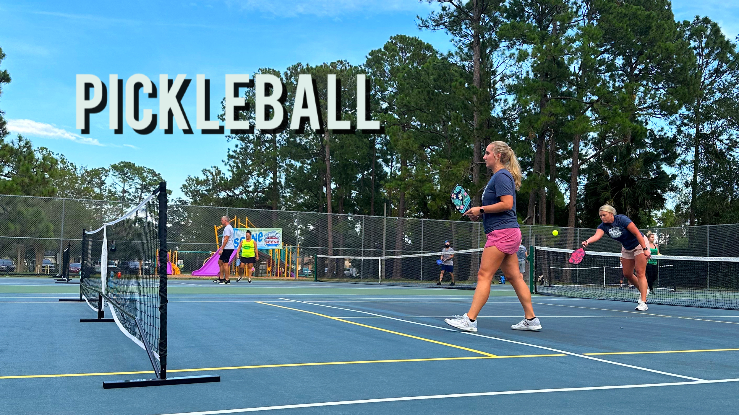 Women playing pickleball on an outdoor court with a slide playground and trees in the background.