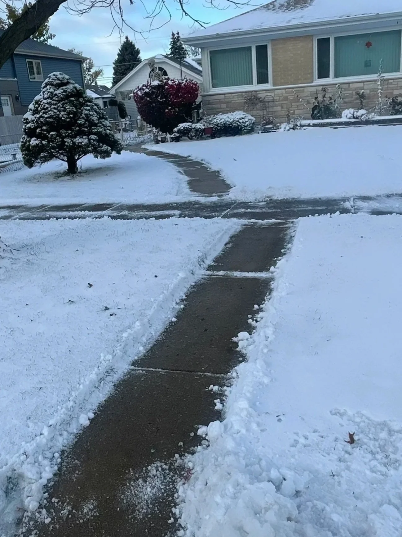 Snow-covered sidewalk and yard in a residential neighborhood, with a house in the background and a tree on the left, during winter.