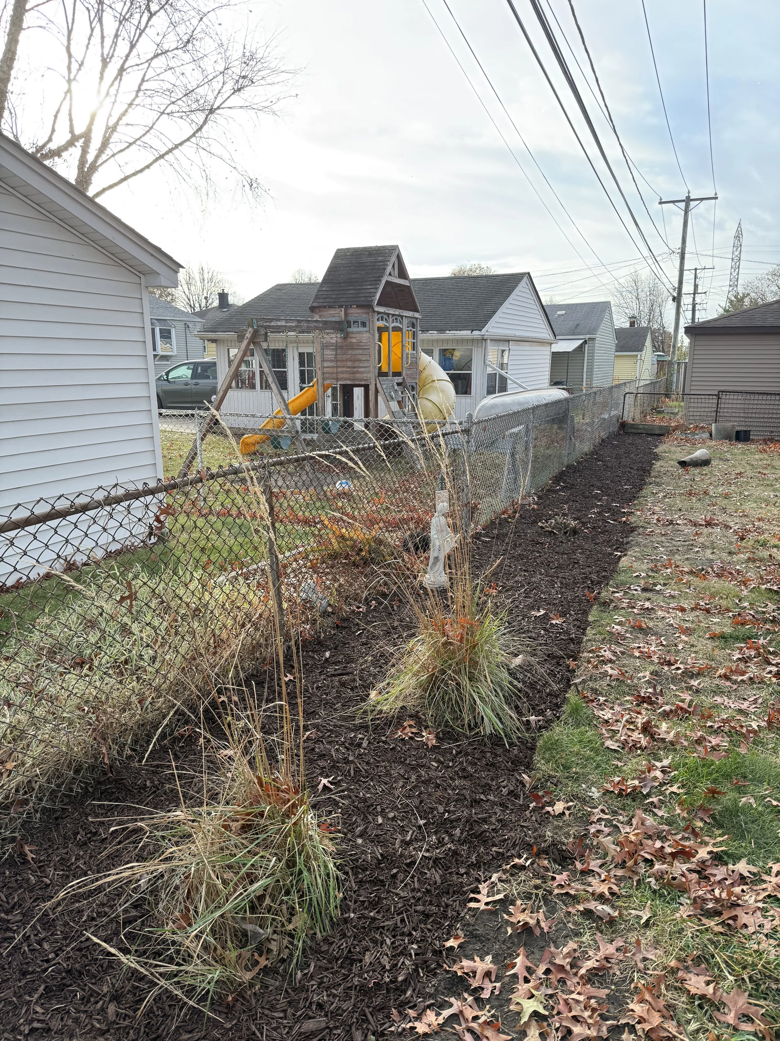 A backyard with a newly mulched flower bed, a chain-link fence, and a children's playhouse with a slide in the background. There are fallen leaves on the grass and a partly cloudy sky.